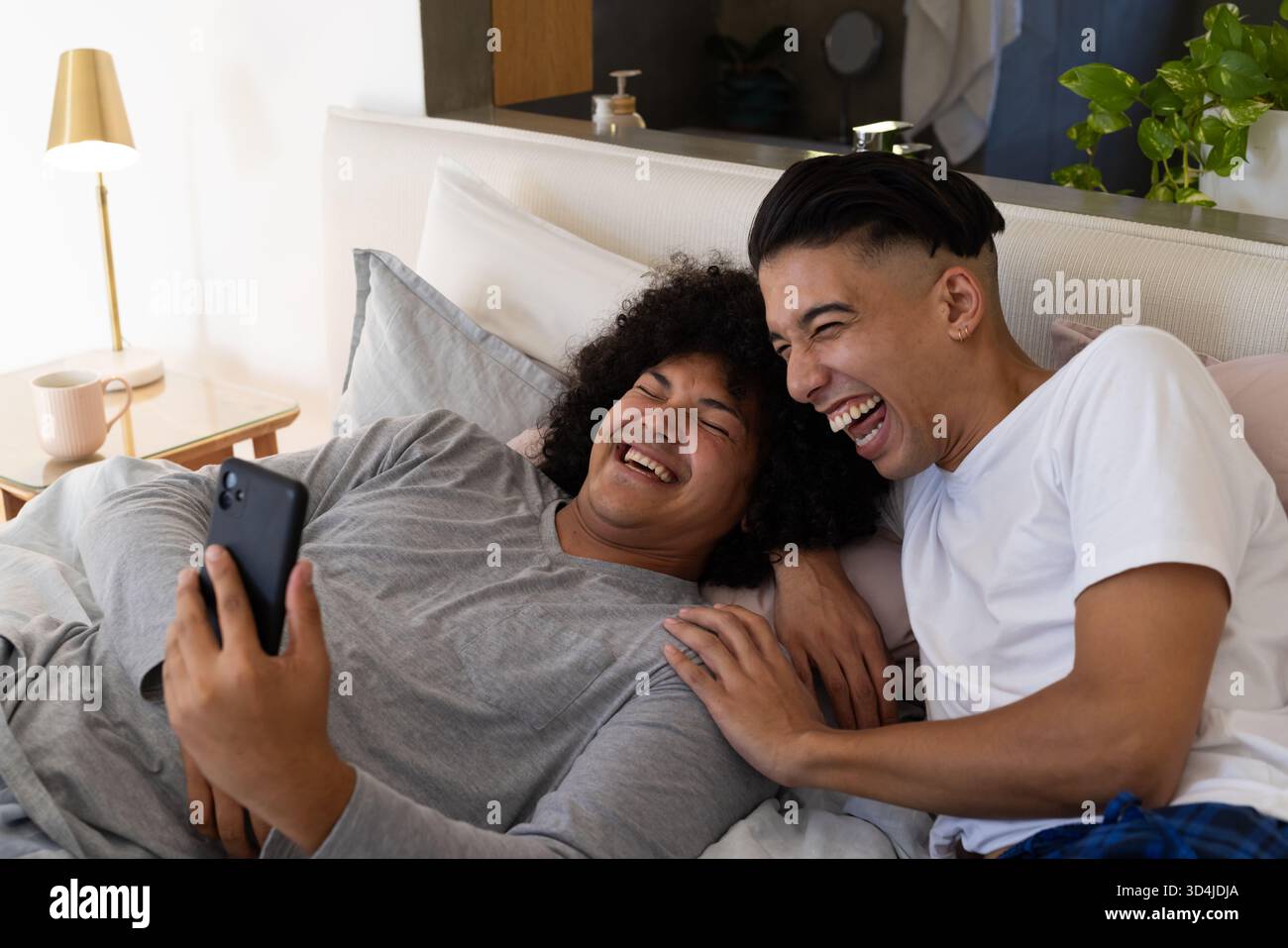 Lachende männliche Freunde, die im modernen Schlafzimmer auf dem Bett liegen, Smartphone benutzen und Becher halten Stockfoto