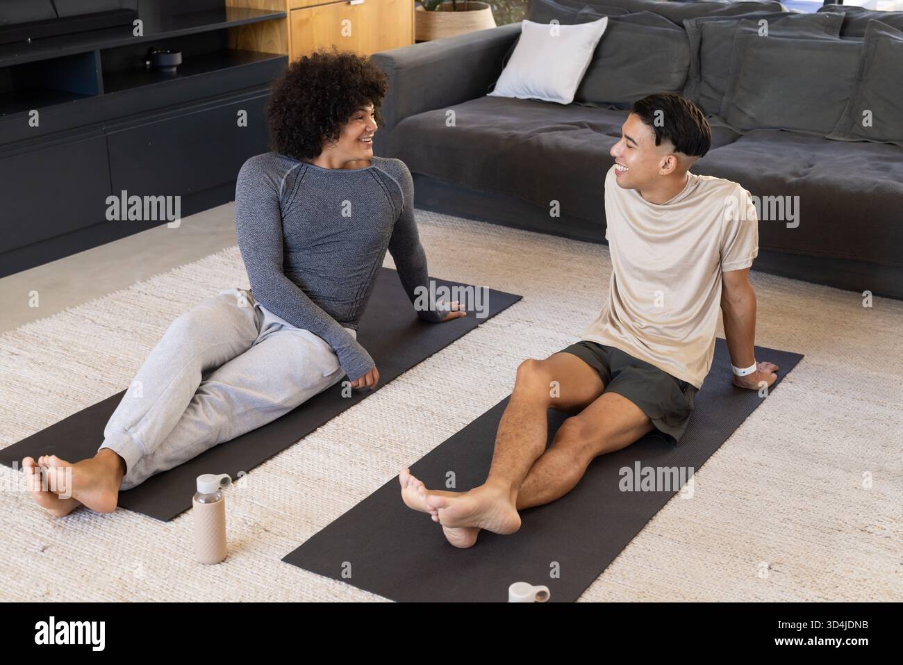 Lachende männliche Freunde sitzen auf Yogamatten im Wohnzimmer, mit Wasserflaschen, moderner Einrichtung Stockfoto