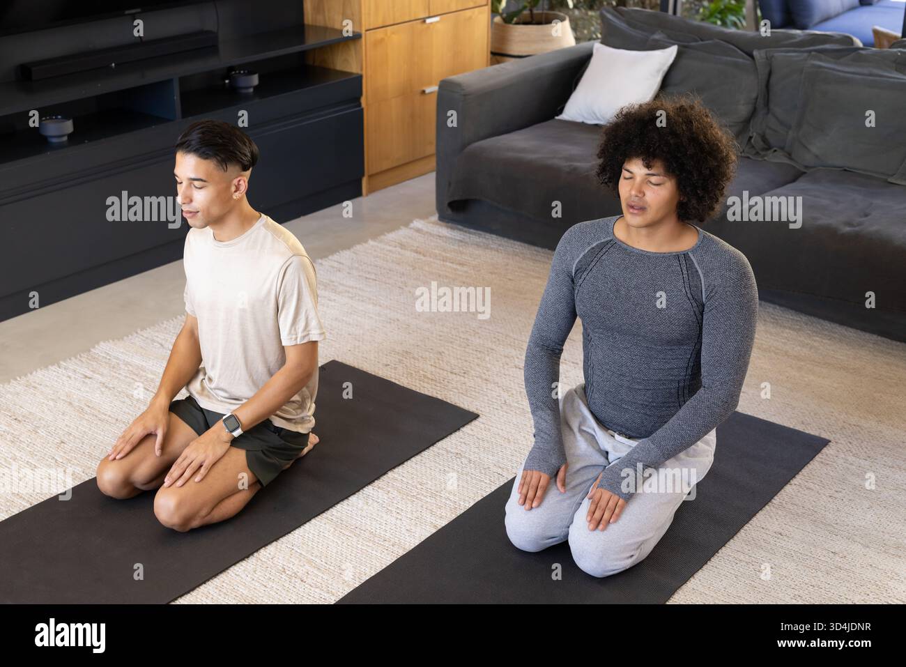 Meditation verschiedener männlicher Freunde, die auf schwarzen Yogamatten im Wohnzimmer knien, mit Topfpflanzen Stockfoto