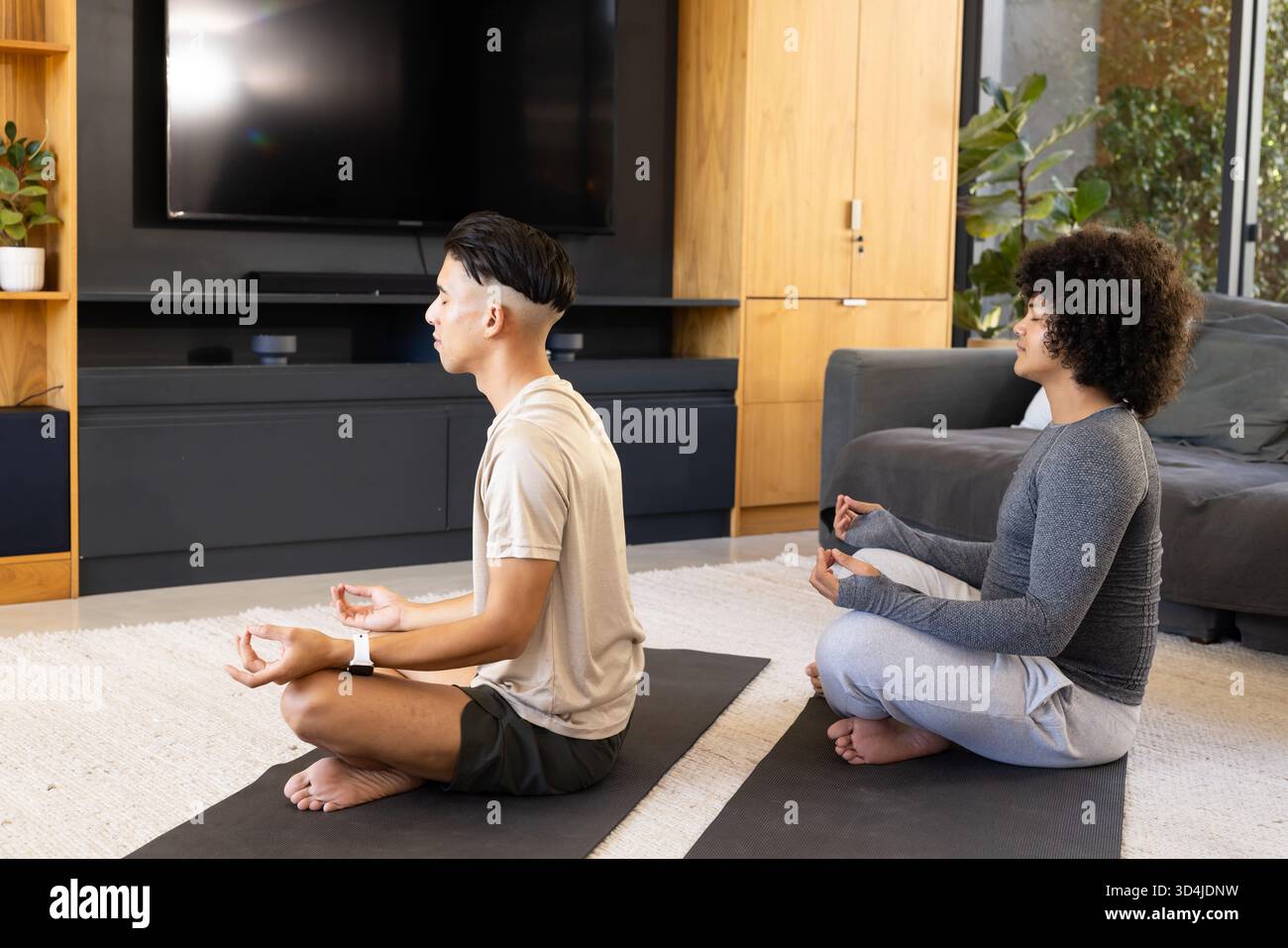 Meditation verschiedener männlicher Freunde im Kreuz im Wohnzimmer, mit Yogamatten und Pflanzen Stockfoto