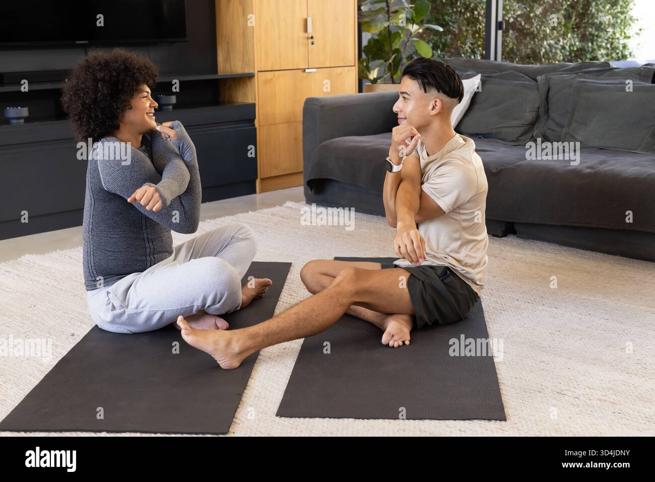 Verschiedene männliche Freunde stehen einander im Wohnzimmer im Kreuz gegenüber, mit schwarzen Yogamatten Stockfoto