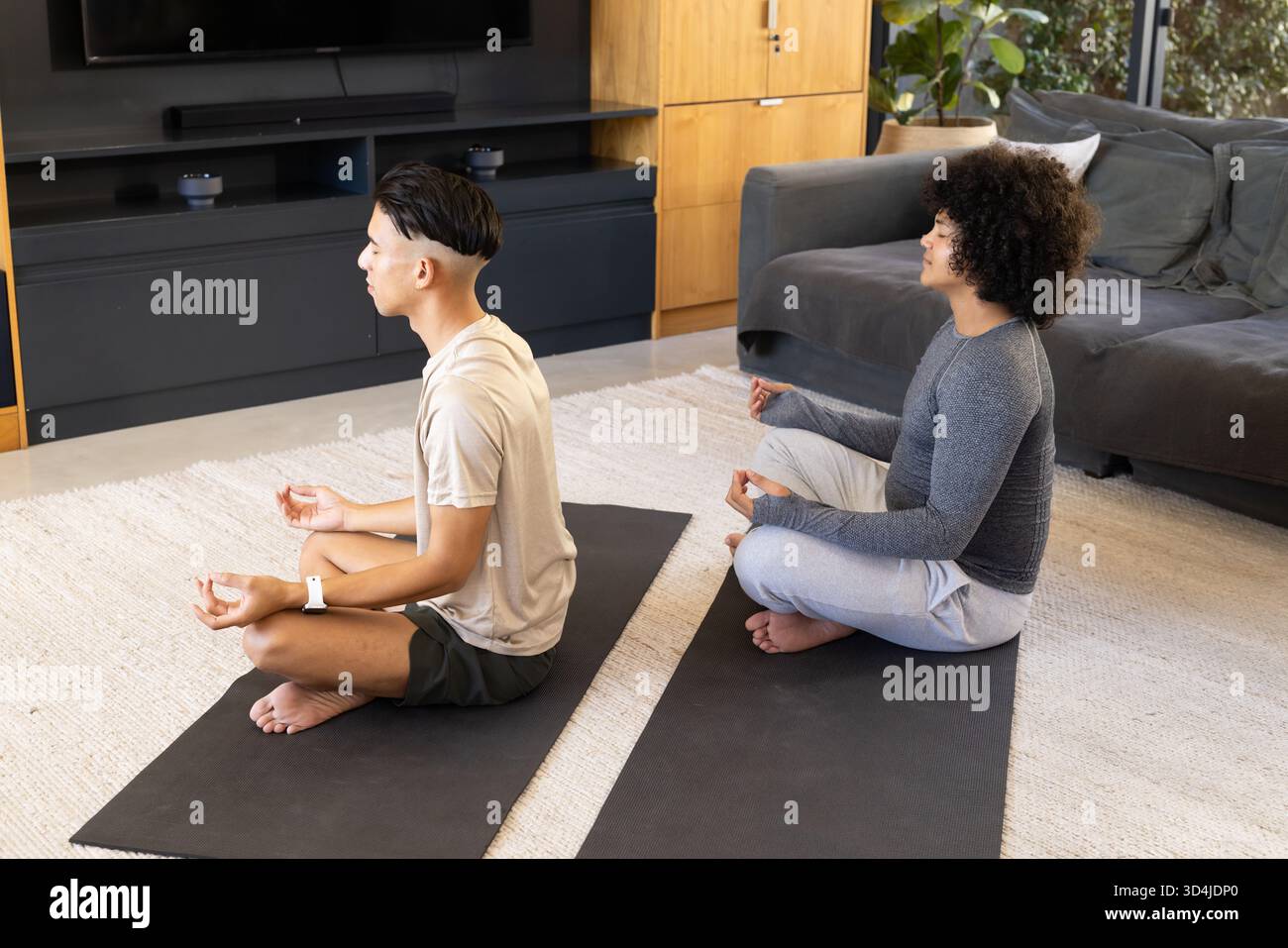 Meditation verschiedener männlicher Freunde, die im Schneidersitz im modernen Wohnzimmer sitzen, auf schwarzen Yogamatten Stockfoto