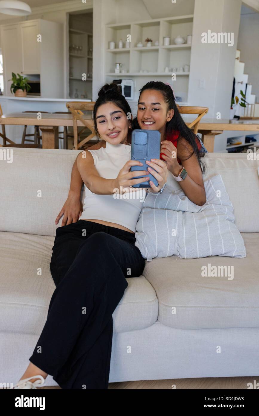 Lächelnde, vielfältige Freundinnen machen Selfie auf hellbeigem Sofa, mit Smartphone und Überwurfkissen Stockfoto