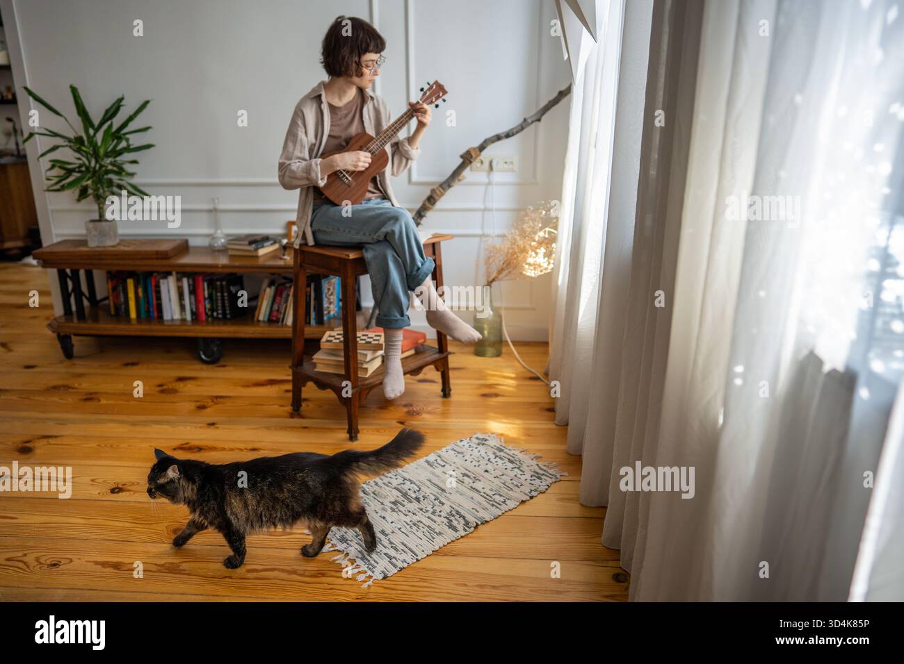 Fokussierte junge Frau, die Ukulele am Fenster einstudiert, eingetaucht in Akkordwiederholung mit Katze herumlaufen Stockfoto