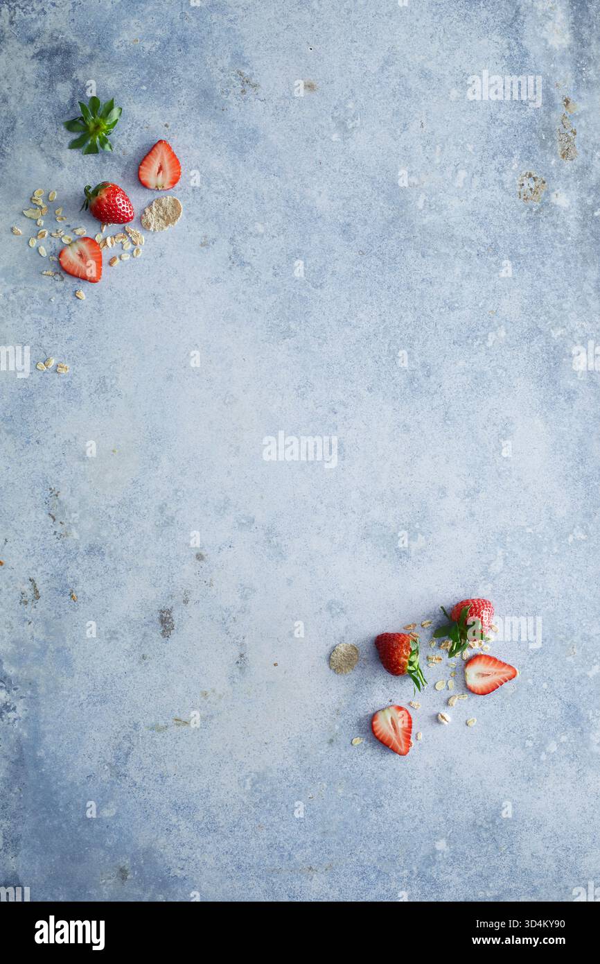 Minimalistische Lebensmittelfotografie von frischen Erdbeeren und verstreuten Müsliflocken auf strukturierter blauer Oberfläche. Perfekt, um gesundes Frühstück zu veranschaulichen Stockfoto
