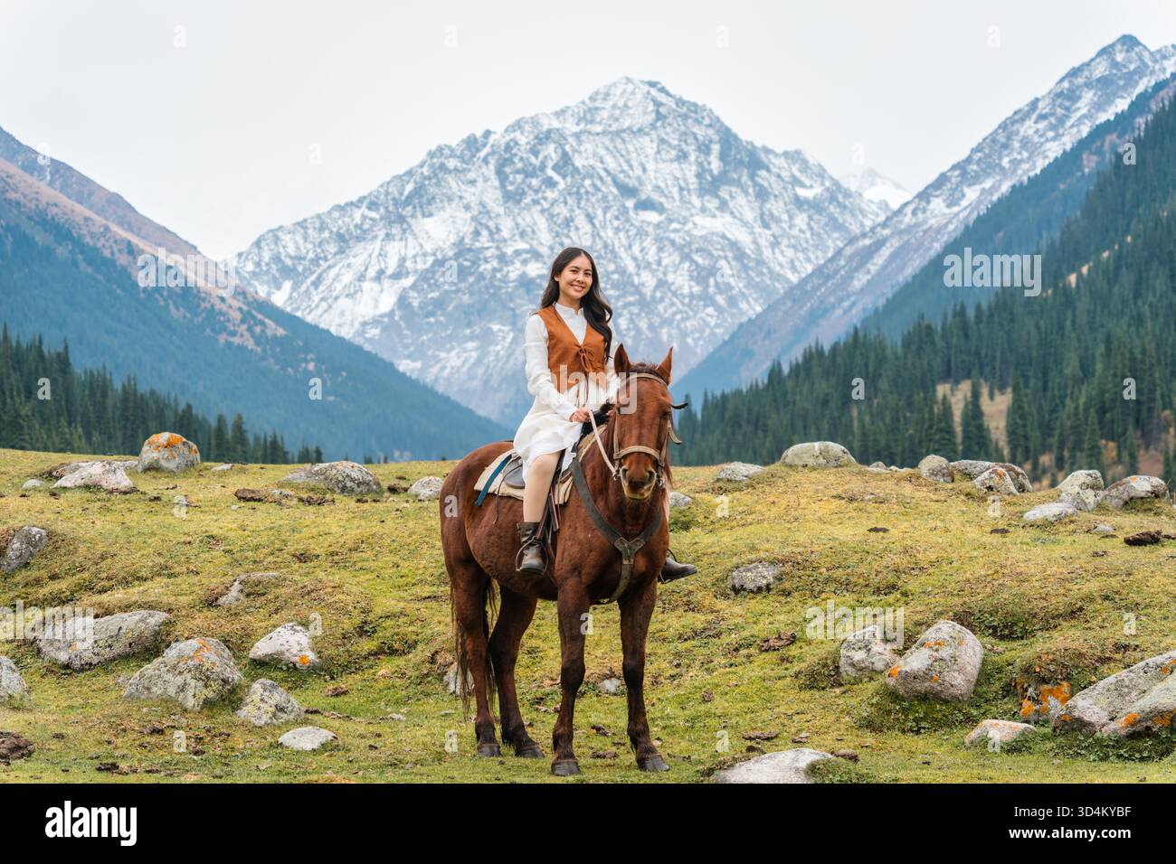 Schöne asiatische Frau reitet Pferd im malerischen Bergtal von Altyn Arashan mit Fluss und schneebedeckten Berg in Kirgisistan Stockfoto
