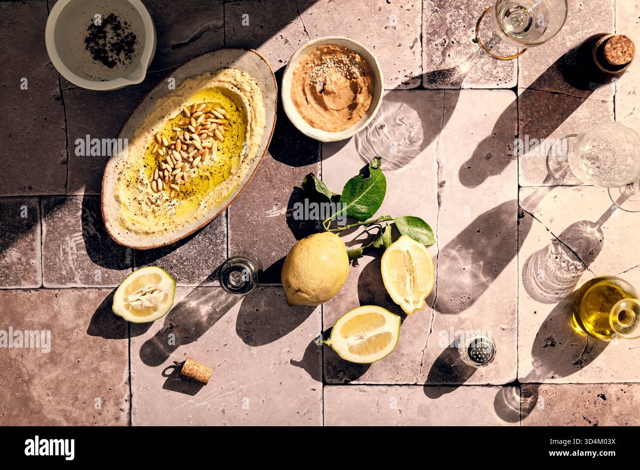 Flacher Hummus mit gerösteten Pinienkernen, Pfeffer und Olivenöl auf einem Teller mit Miso- und Sesamsamen, frischen Zitronen und Weingläsern in hartem Lig Stockfoto