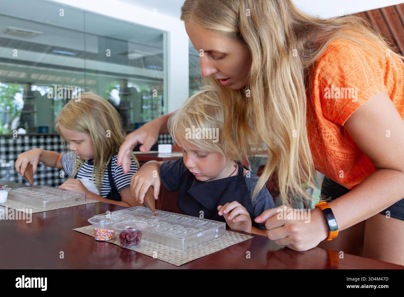 Kinder und ihre Mutter nehmen an einem Workshop zur Schokoladenherstellung in Bali teil, bei dem Süßigkeiten mit Belag dekoriert werden. Eine unterhaltsame und lehrreiche Familienaktivität Stockfoto