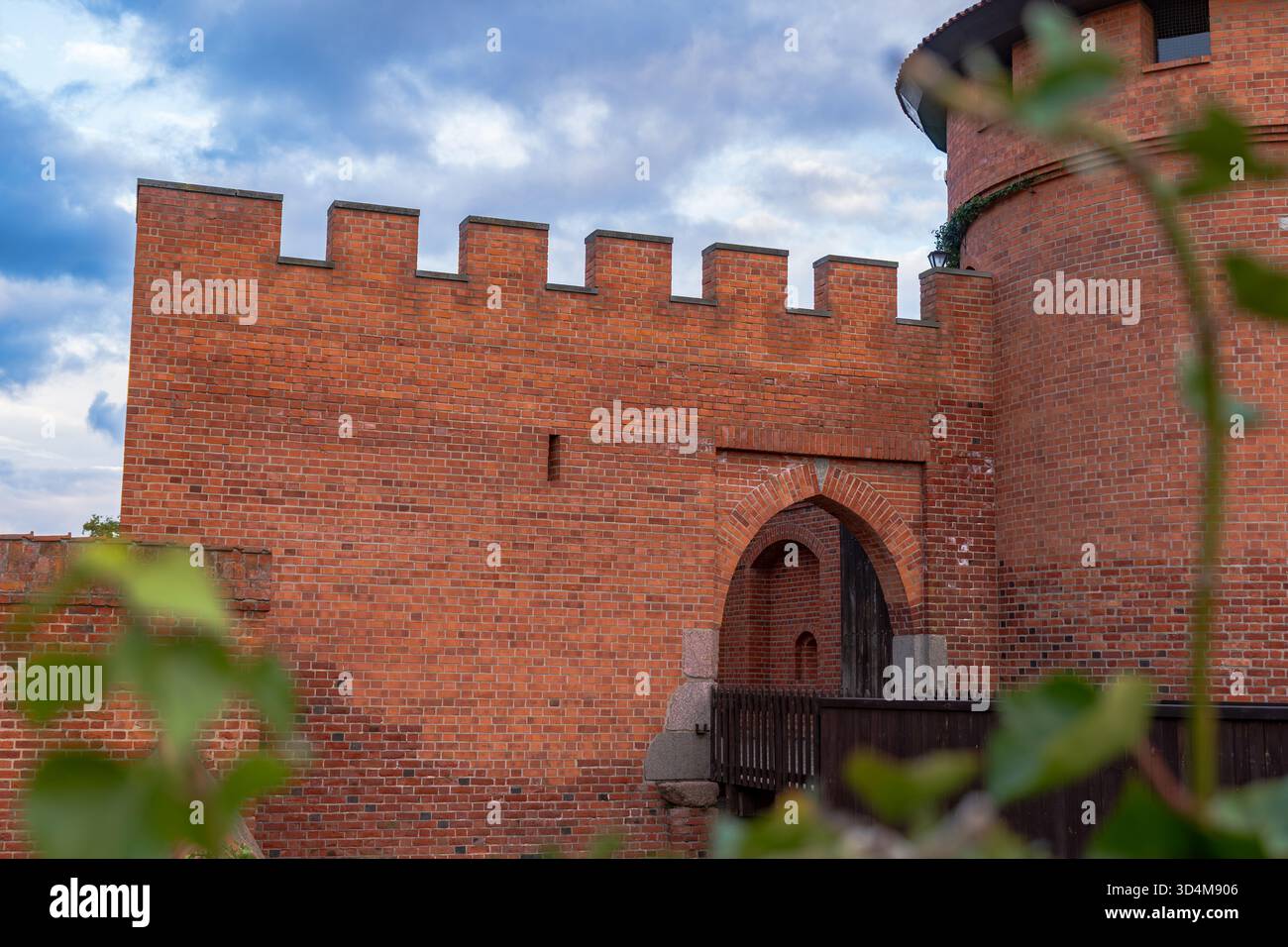 Aus nächster Nähe sehen Sie eine zinnenförmige Mauer, ein bogenförmiges Tor und einen runden Turm auf der Burg Malbork, Polen. Warmes Licht am späten Tag offenbart Ziegelstruktur, Pfeilschlitze, Stockfoto
