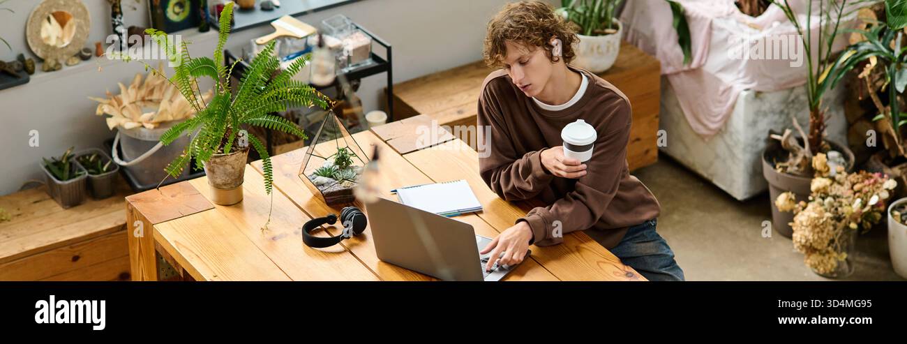 Ein junger Mann greift mit seinem Laptop in Berührung, während er einen Kaffee in einer künstlerischen Umgebung genießt. Stockfoto