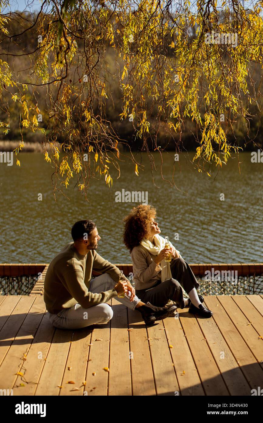 Ein Paar entspannt sich am Ufer und schlürft warme Getränke, während die Sonne durch die Bäume scheint. Die Atmosphäre fühlt sich friedlich und intim an. Stockfoto