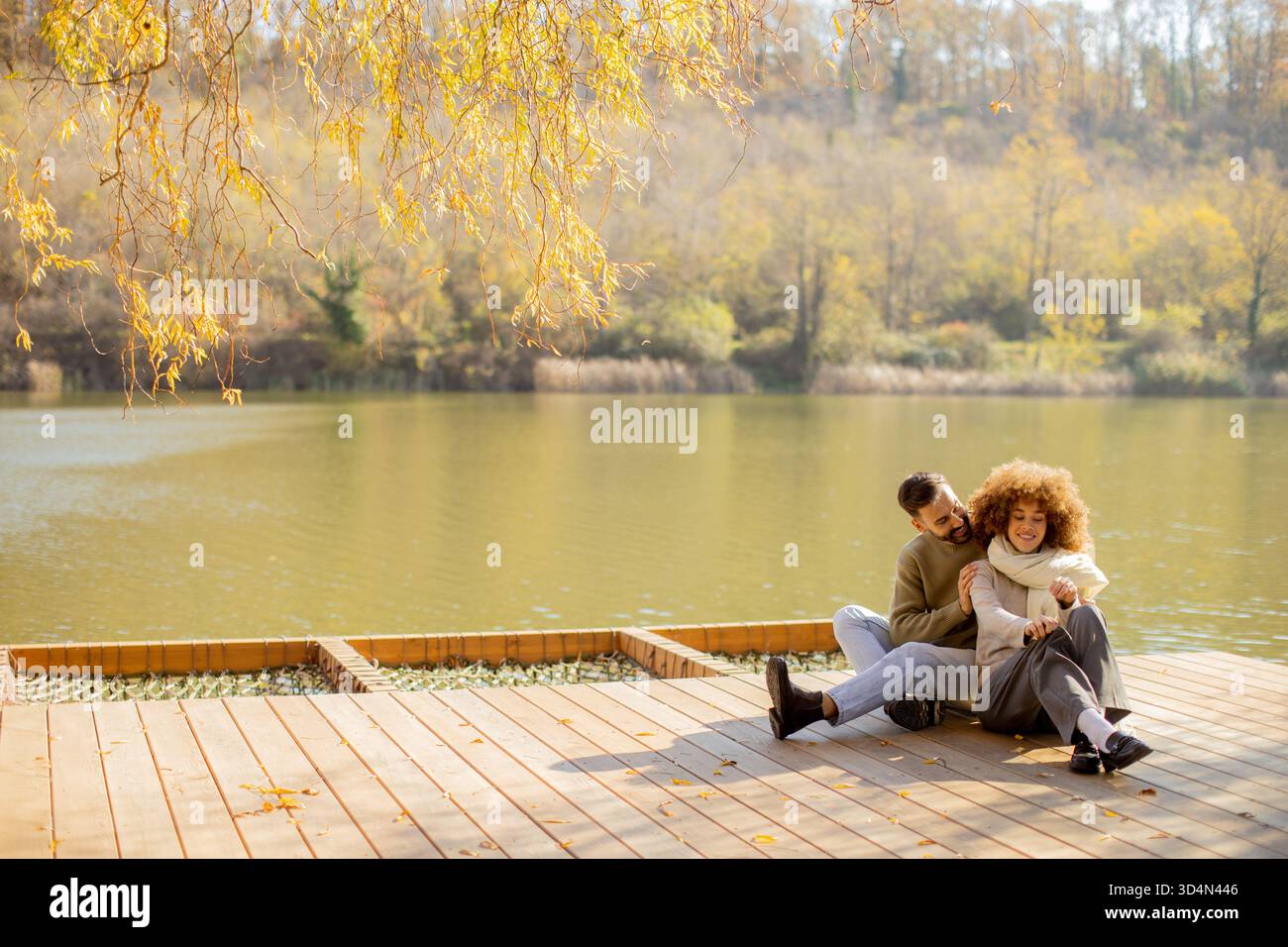 Zwei Personen sitzen bequem an einem ruhigen Seeufer und teilen sich Lachen und Wärme in leuchtenden Herbstfarben. Stockfoto