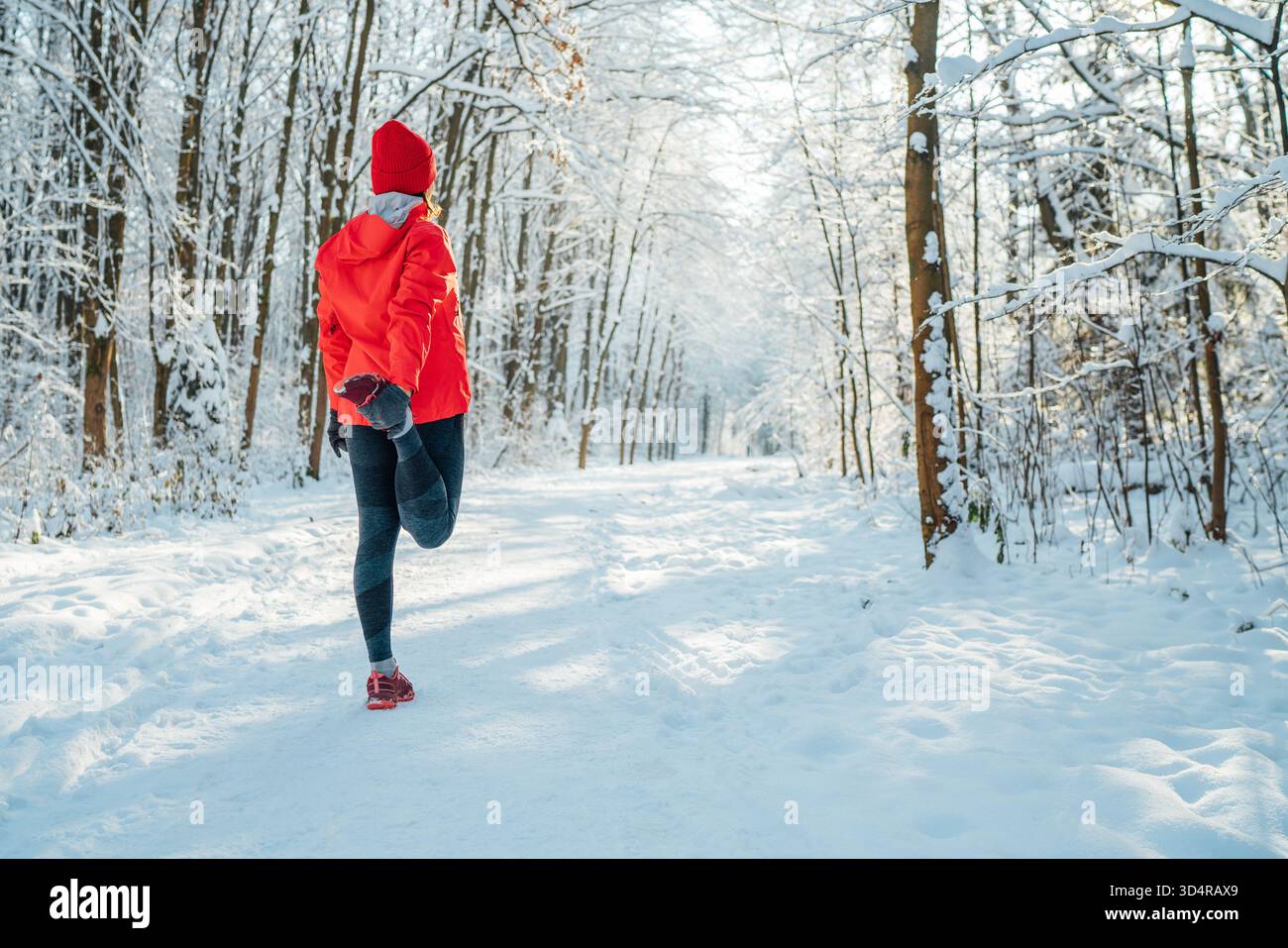 Trailläuferin in leuchtend roter, winddichter Jacke wärmt sich auf, während sie an einem sonnigen frostigen Tag auf dem malerischen verschneiten Waldweg unterwegs ist. Sportliche Klimaanlage Stockfoto