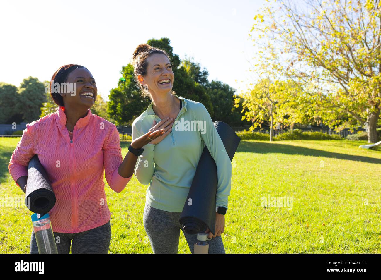 Lächelnde, vielfältige Freundinnen, die sich im Park zum Workout ausstrecken, mit Yogamatten und Wasserflaschen Stockfoto