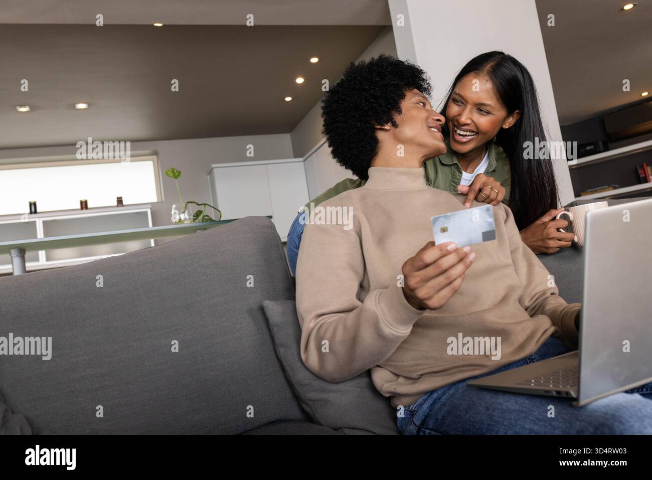 Stöbern Sie auf einem Laptop auf einem grauen Sofa im modernen Wohnzimmer mit Kreditkarte Stockfoto