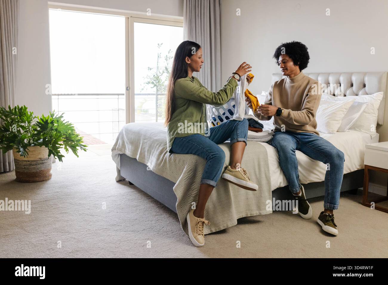 Verschiedene Sortiersachen für Paare auf Queen-Size-Bett Rand in modernem Schlafzimmer, mit weißem Wäschekorb Stockfoto