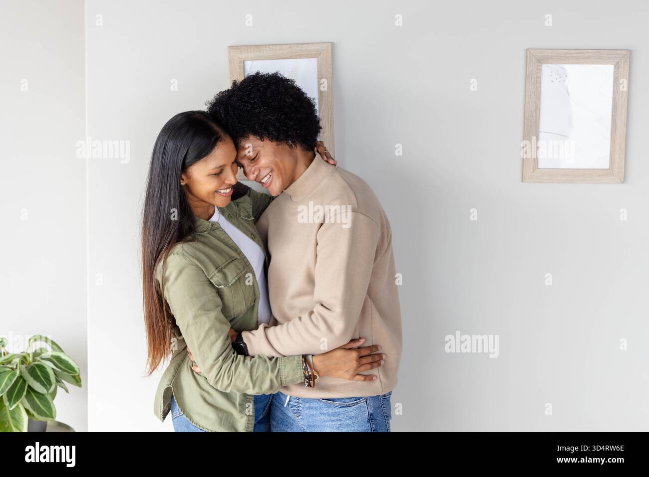 Umarmtes, vielseitiges Paar, das sich nahe in einem hellen Wohnzimmer mit hölzernen Bilderrahmen und Pflanzen lehnt Stockfoto