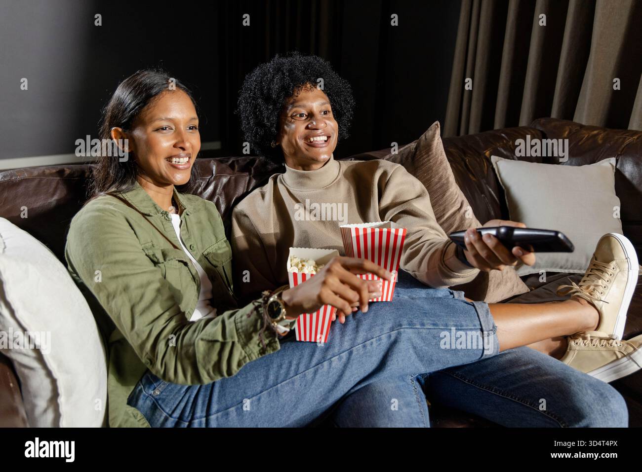 Sie teilen Popcorn mit verschiedenen Freunden, die auf einer Ledercouch in schwach beleuchtetem Wohnzimmer sitzen, mit TV-Fernbedienung Stockfoto