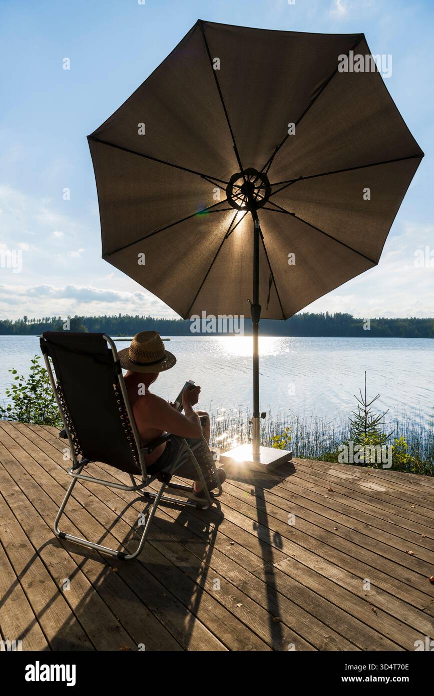 Der Rücken eines älteren Mannes, der in einem Liegestuhl auf einer Holzterrasse am See sitzt Stockfoto
