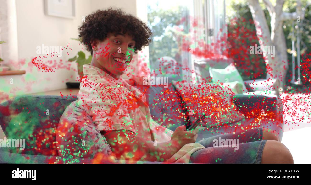 Lächelnder Erwachsener, der eine Jeansshorts trägt, hält das Telefon im Wohnzimmer auf dem Sofa, mit Konfetti-Überzug Stockfoto