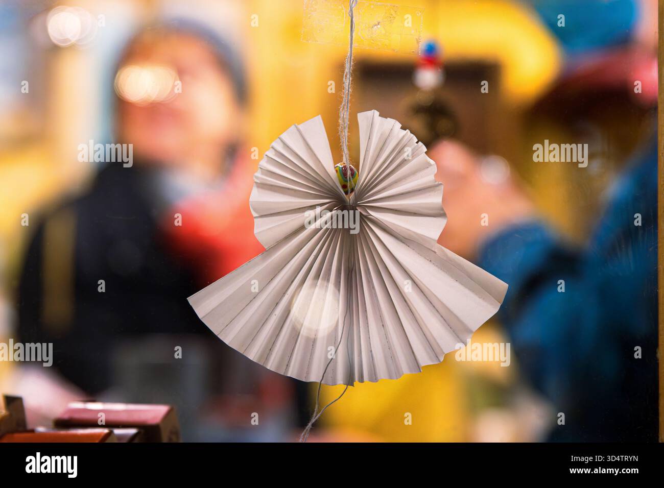 Handgefertigtes EngelsOrigami-Ornament mit verschwommenem festlichem Weihnachtsmarkt und Menschen im Hintergrund. Unterstreicht den Urlaubsgeist durch DIY-Dekoraten Stockfoto