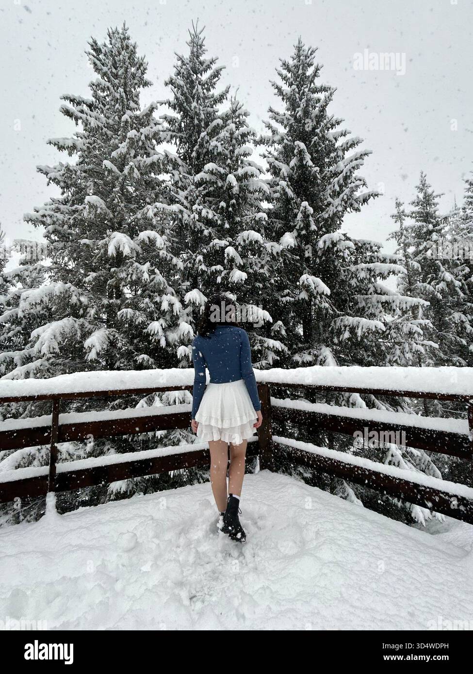 Lifestyle-Szene im Winter mit einer jungen Frau, die auf einer Holzterrasse in einem verschneiten Bergwald starken Schneefall genießt. Perfekt für Reisen, Mode, Urlaub - Smartphone-aufgenommenes Stockfoto