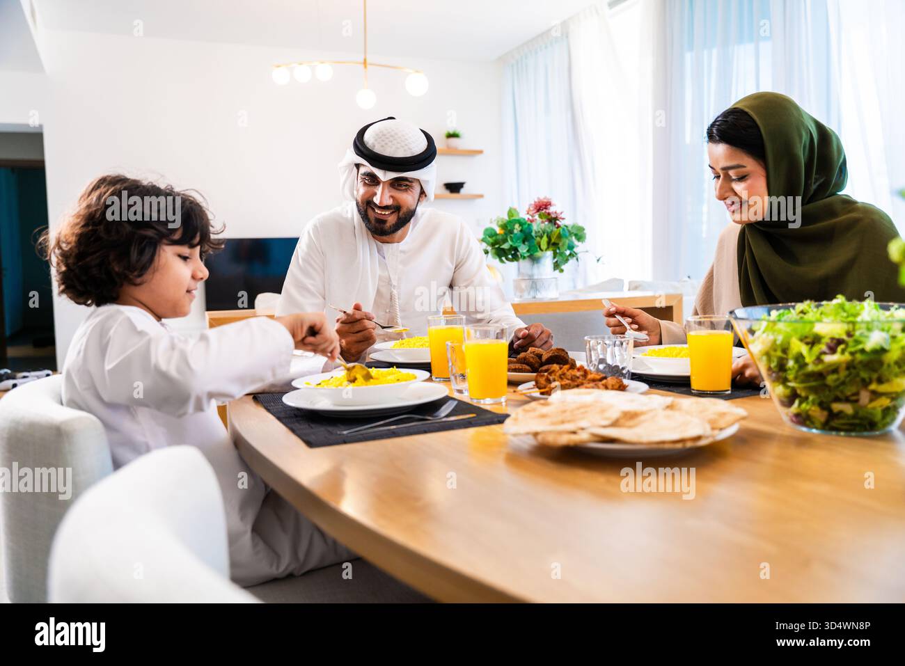 Familie aus dem Nahen Osten trägt traditionelle emiratische Kleidung und verbringt Zeit zu Hause zusammen. Familie aus dem Nahen Osten, die ihr Mittagessen am Esstisch im Inneren genießt Stockfoto