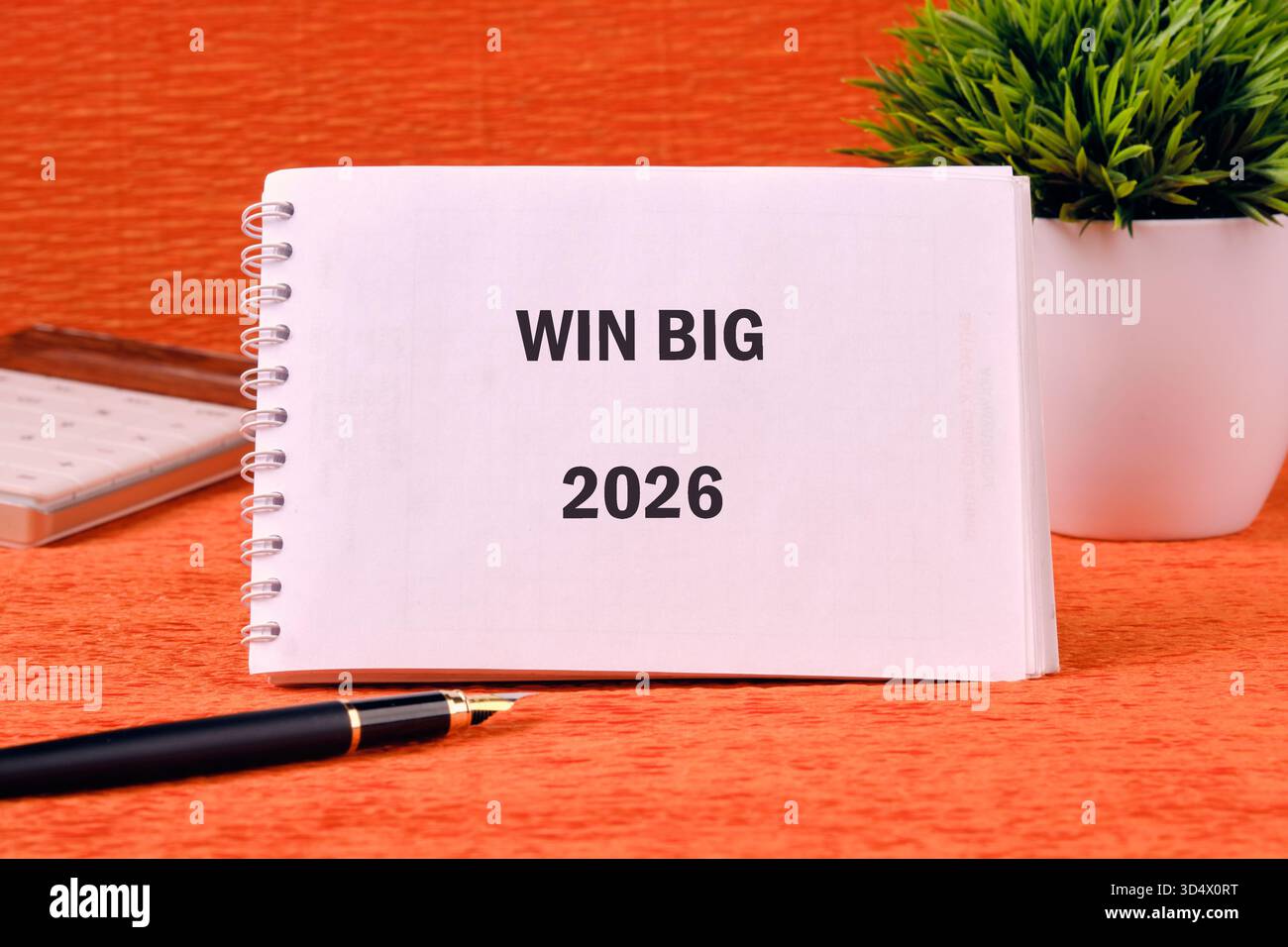 Ein spiralförmiges Notizbuch mit Win Big 2026 liegt auf einer gemusterten orangefarbenen Oberfläche, begleitet von einem Stift, einem Taschenrechner und einer kleinen grünen Pflanze in einem weißen Topf. Stockfoto