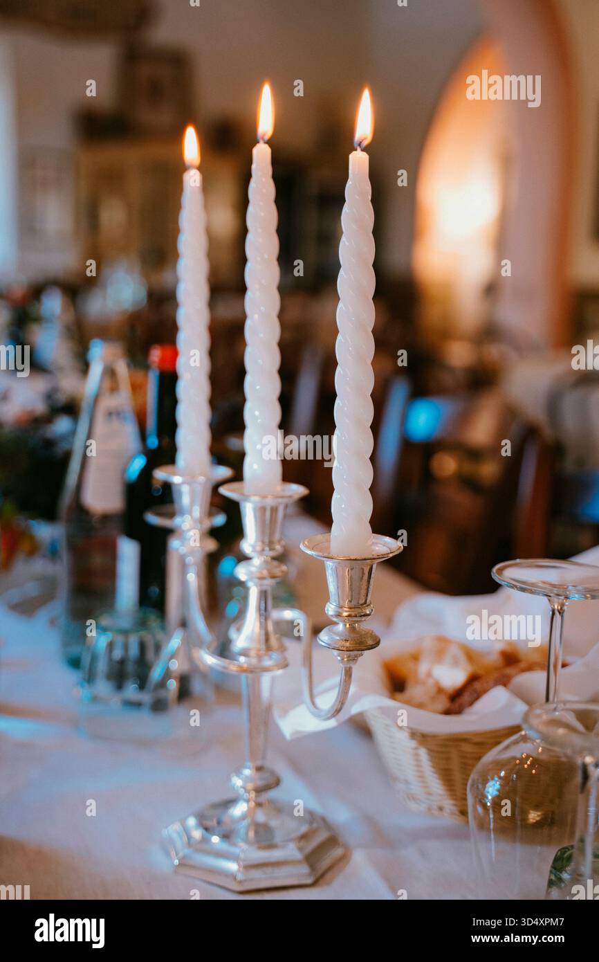 Drei elegante verdrehte Kerzen brennen hell in silbernen Kerzenleuchtern auf einem wunderschön gestalteten Esstisch, der für ein warmes Treffen mit Brot und Glas eingerichtet ist Stockfoto