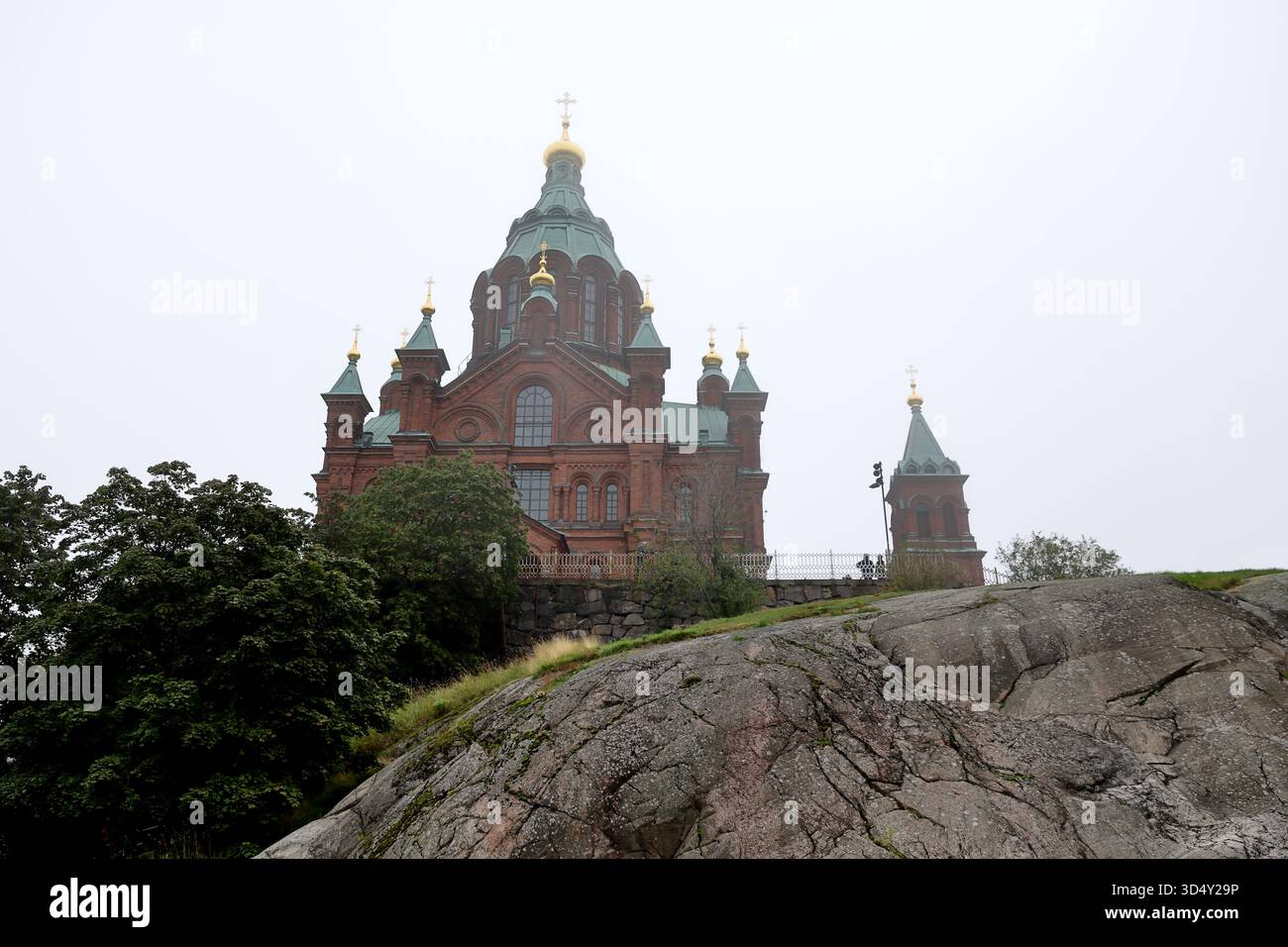 Die Kathedrale von Uspenski erhebt sich über Granitgestein und Bäume in Helsinki, Finnland, die von unten an einem nebeligen Tag mit diffusem Licht und hellem Himmel fotografiert wurde. Stockfoto