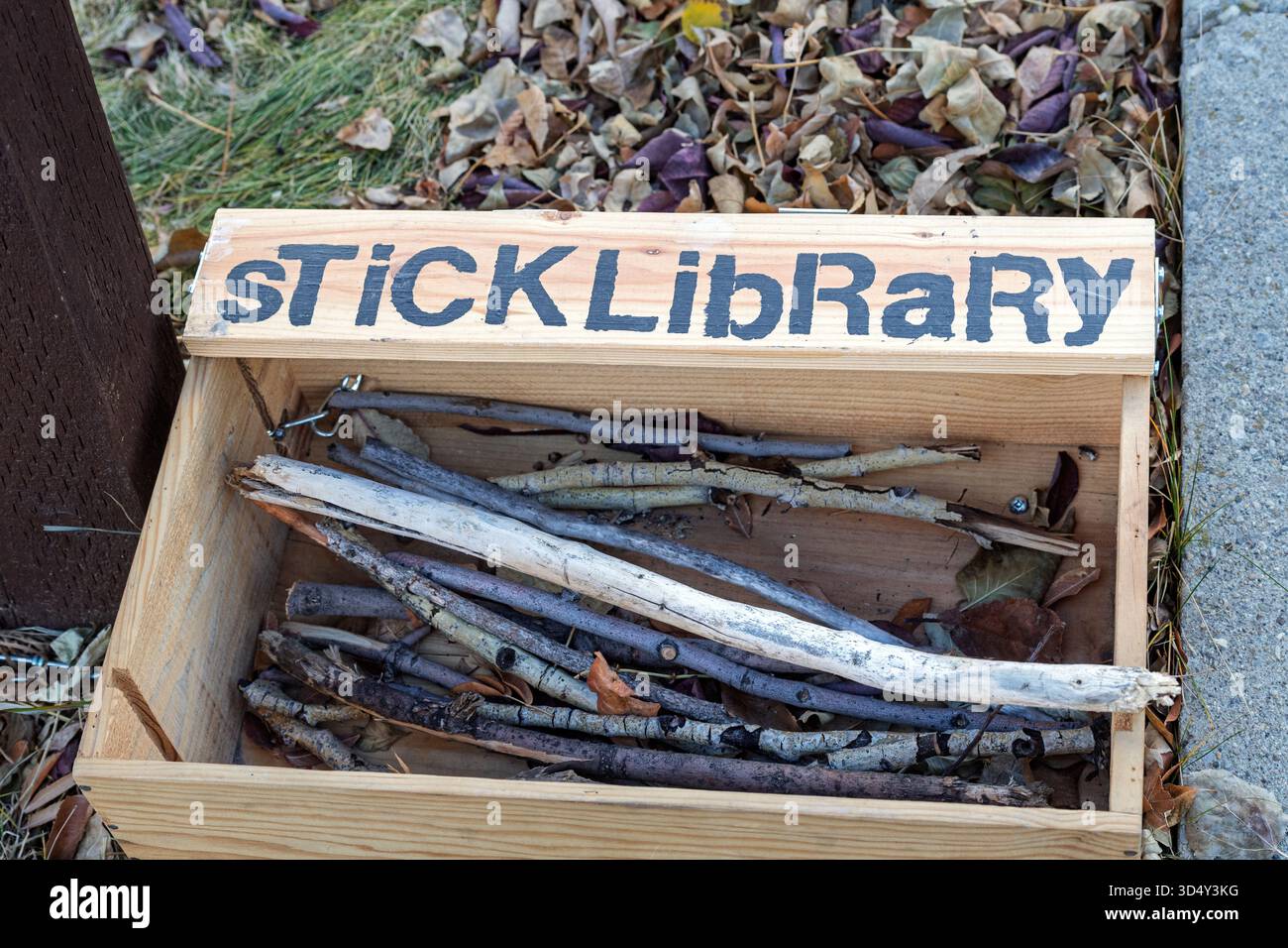 Eine Holzkiste mit der Aufschrift „Stick Library“ steht im Freien, gefüllt mit kleinen Stäben. Das verspielte Schild lädt Menschen oder Hunde zum Ausleihen und Rückgeben von Stäben ein Stockfoto