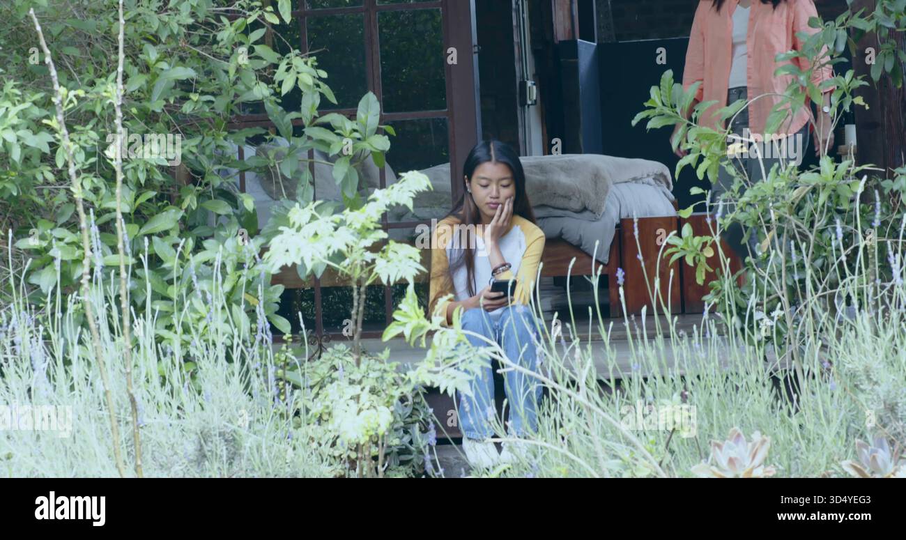 Sitzende Erwachsene asiatische Frau, die ihr Smartphone auf hölzernen Terrassentreppen blättert, mit gelbem Oberteil und Jeans Stockfoto