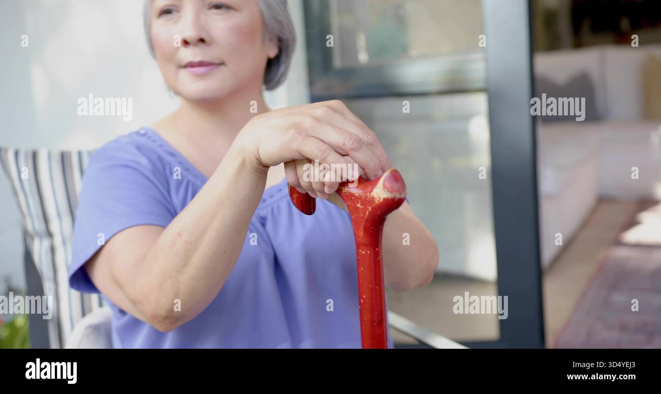 Sitzende ältere asiatische Frau mit Lavendeloberteil, die roten Stock auf der Veranda mit gestreiftem Kissen hält Stockfoto