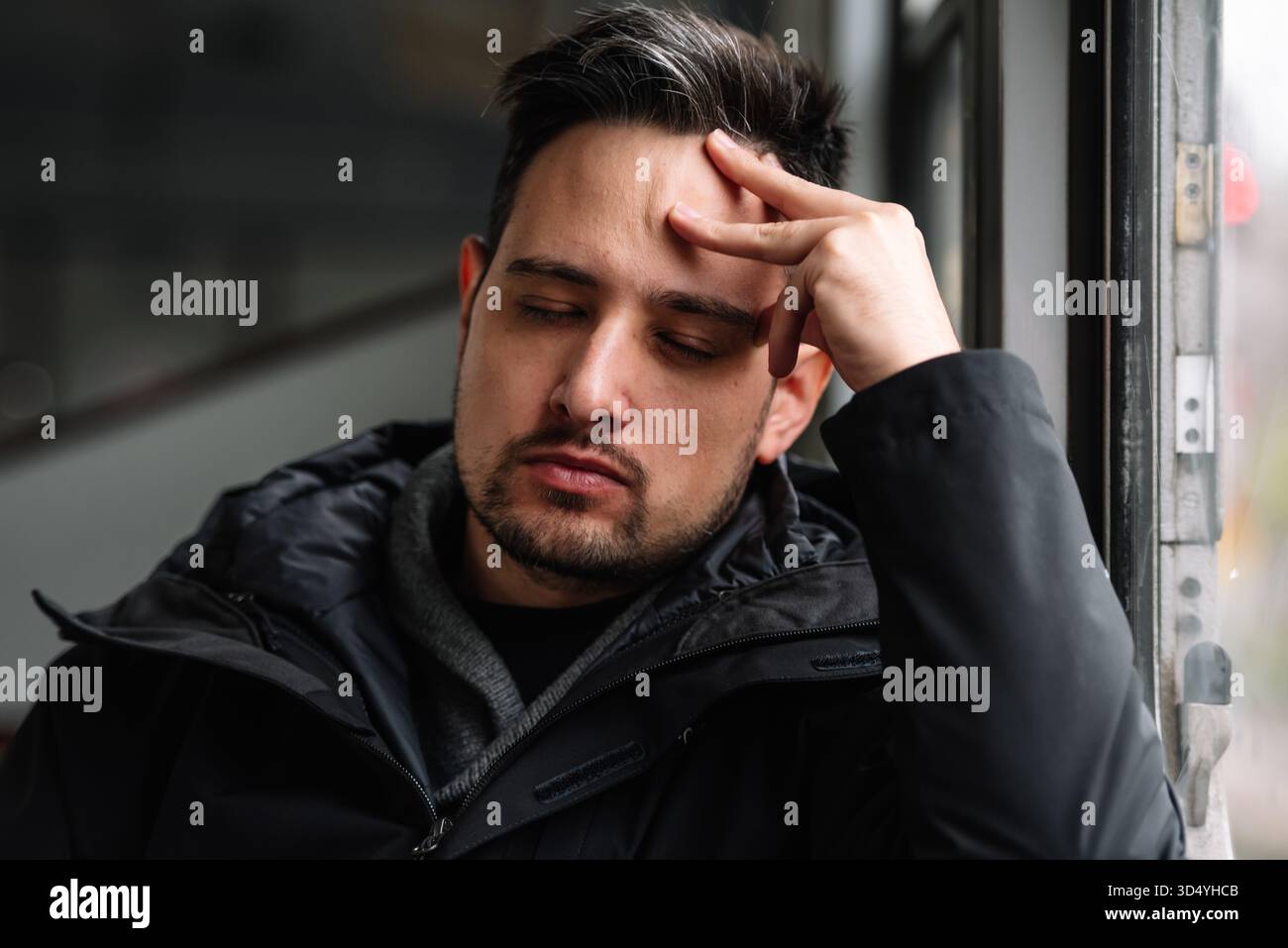 Müder junger Mann mit geschlossenen Augen, der Kopf auf der Hand am Fenster liegt Stockfoto
