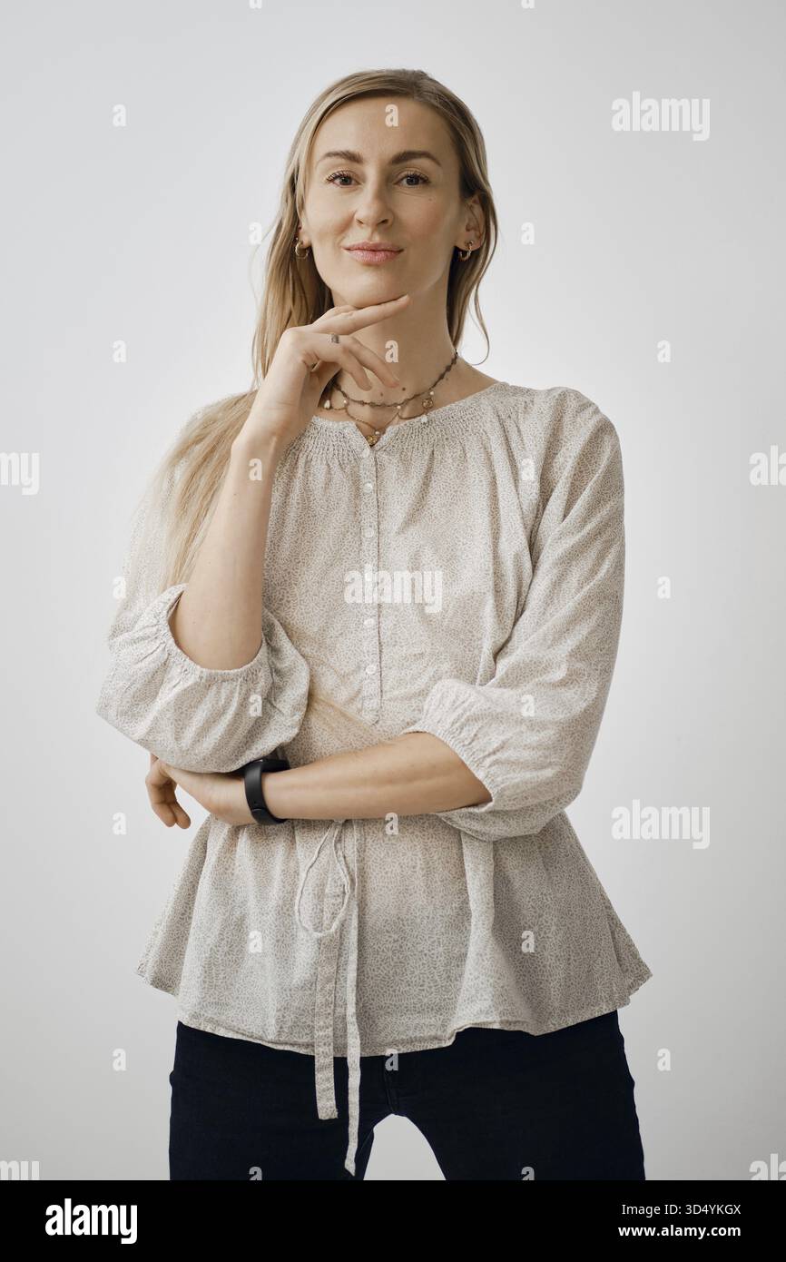 Eine Frau steht in entspannter Pose mit einer Hand am Kinn und trägt eine helle, lässige Bluse. Sie sieht entspannt aus, mit einem leichten Lächeln Stockfoto