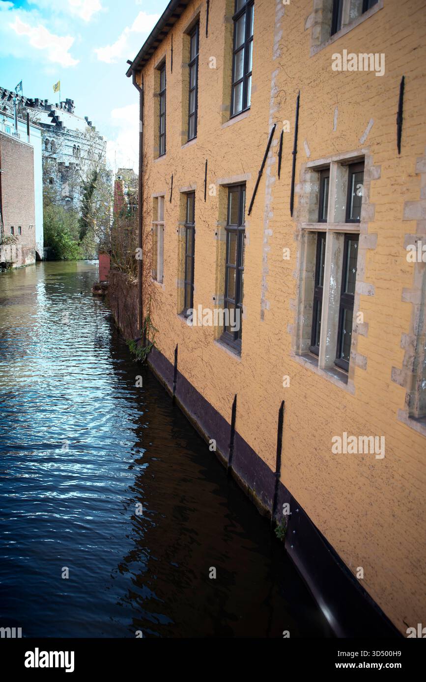 Ein gelbes Gebäude am Fluss mit hohen Fenstern steht neben einem ruhigen Kanal in Gent, Belgien, das das Sonnenlicht reflektiert, und das Schloss Gravensteen ist im Th sichtbar Stockfoto