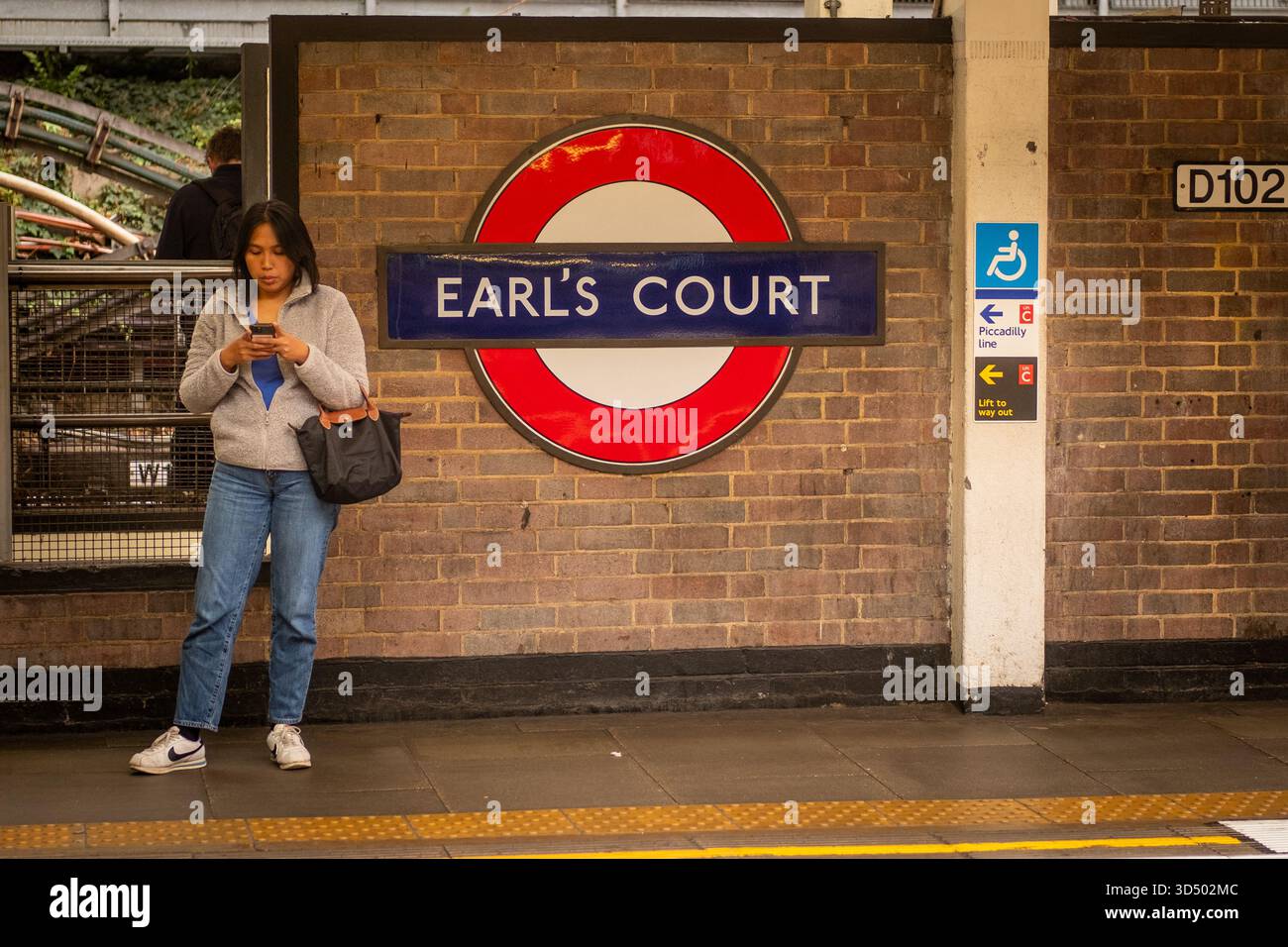 LONDON – 15. OKTOBER 2025: Junge Frau nutzt das Telefon an der Earl’s Court Station Platform, District & Piccadilly Line Stockfoto