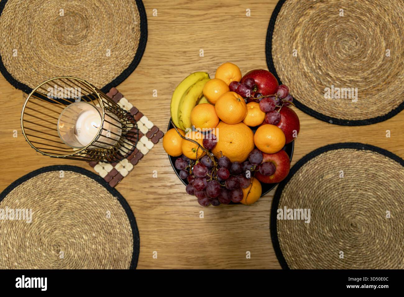 Farbenfrohe Obstschale und Kerze auf dem Esstisch. Blick von oben auf eine lebendige Obstschale mit Bananen, Trauben, Äpfeln und Orangen neben einer beleuchteten Kerze auf einem Stockfoto
