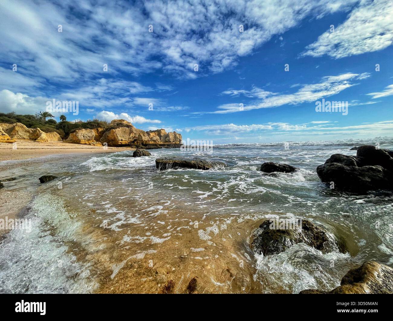 Schaumige Wellen krachen auf den Felsen der abgelegenen Praia dos Bés, Albufeira, Algarve, Portugal, unter Klippen und einem lebhaften Himmel voller Wolken Stockfoto