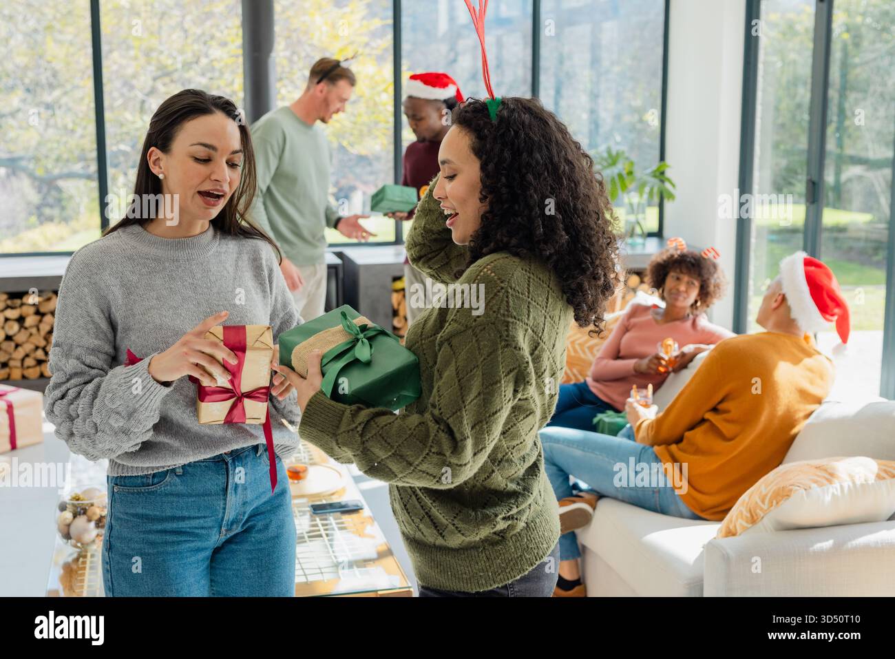 Verschiedene weibliche Kollegen, die ein Stirnband tragen, tauschen im Wohnzimmer mit Glastisch umwickelte Geschenke aus Stockfoto