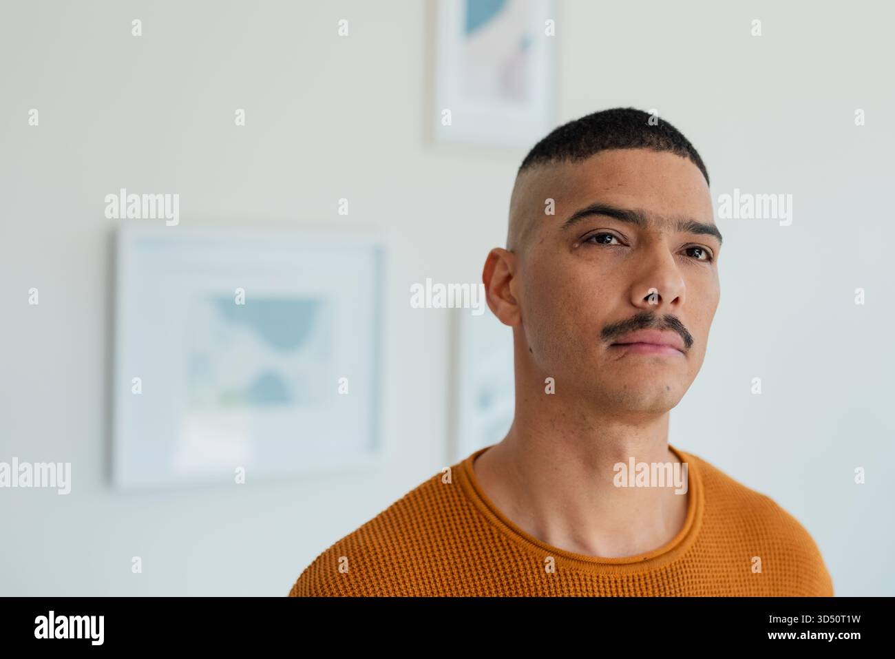 Mann mit orangefarbenem Pullover, der zu Hause an einer hellen Wand steht, mit abstrakten Kunstdrucken, Kopierraum Stockfoto