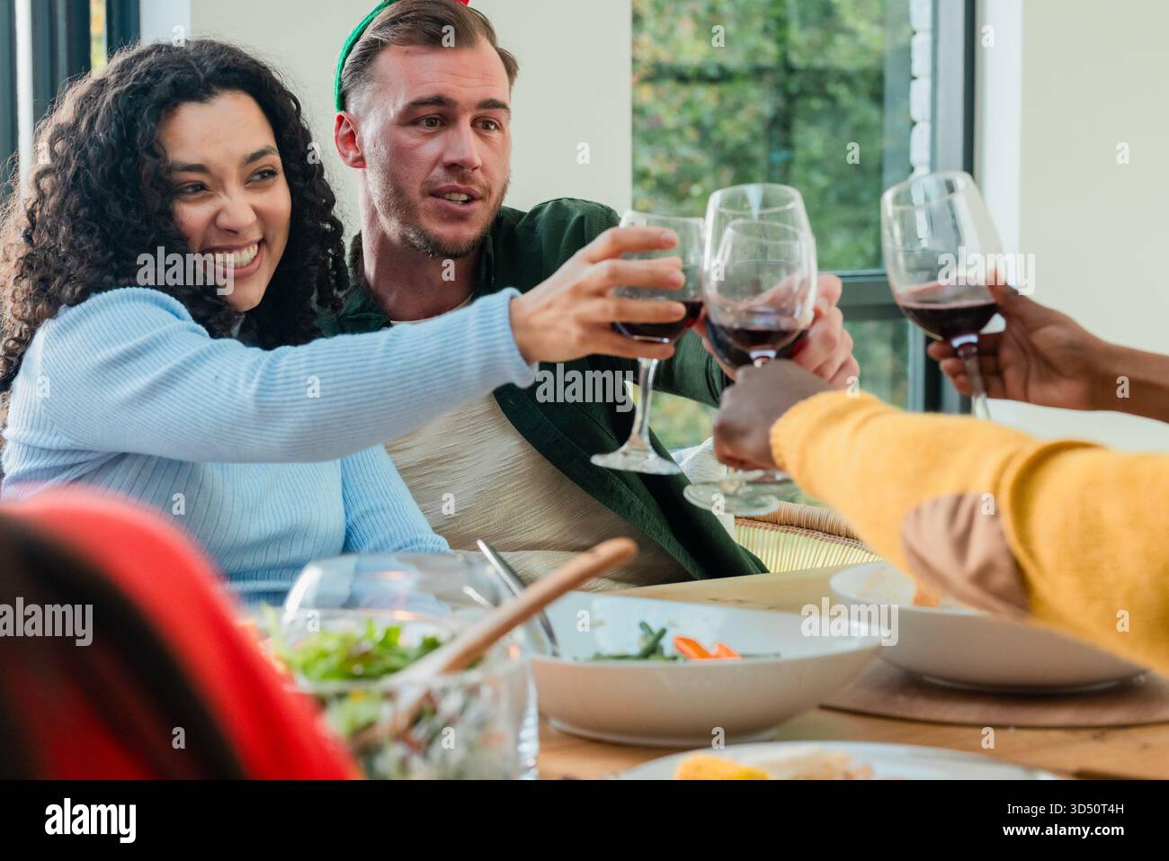Verschiedene Freunde klirren Rotweingläser, die am Esstisch am Fenster mit Blick auf den Garten sitzen Stockfoto