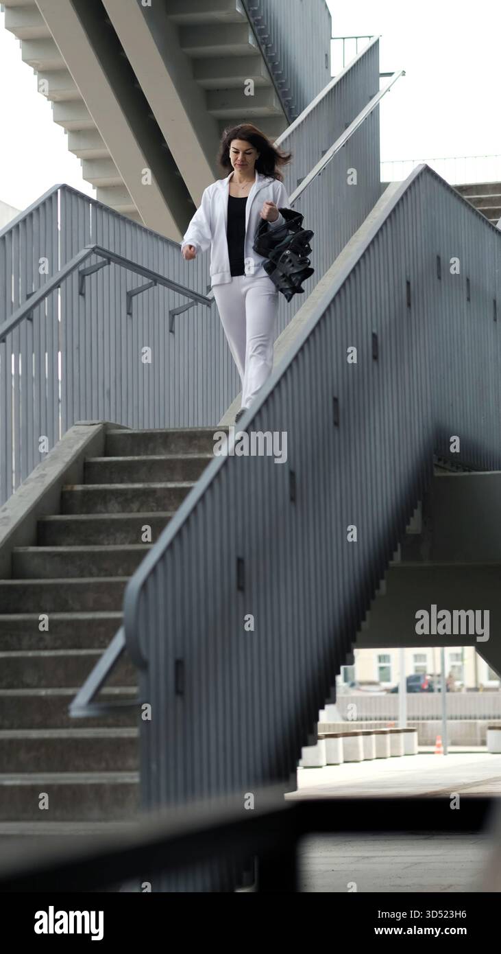 Eine selbstbewusste Frau in weißer Sportswear geht eine städtische Treppe hinunter und hält Kangoo-Schuhe in der Hand. Die strukturierten Linien der Treppe bilden den Rahmen für das Thema Stockfoto