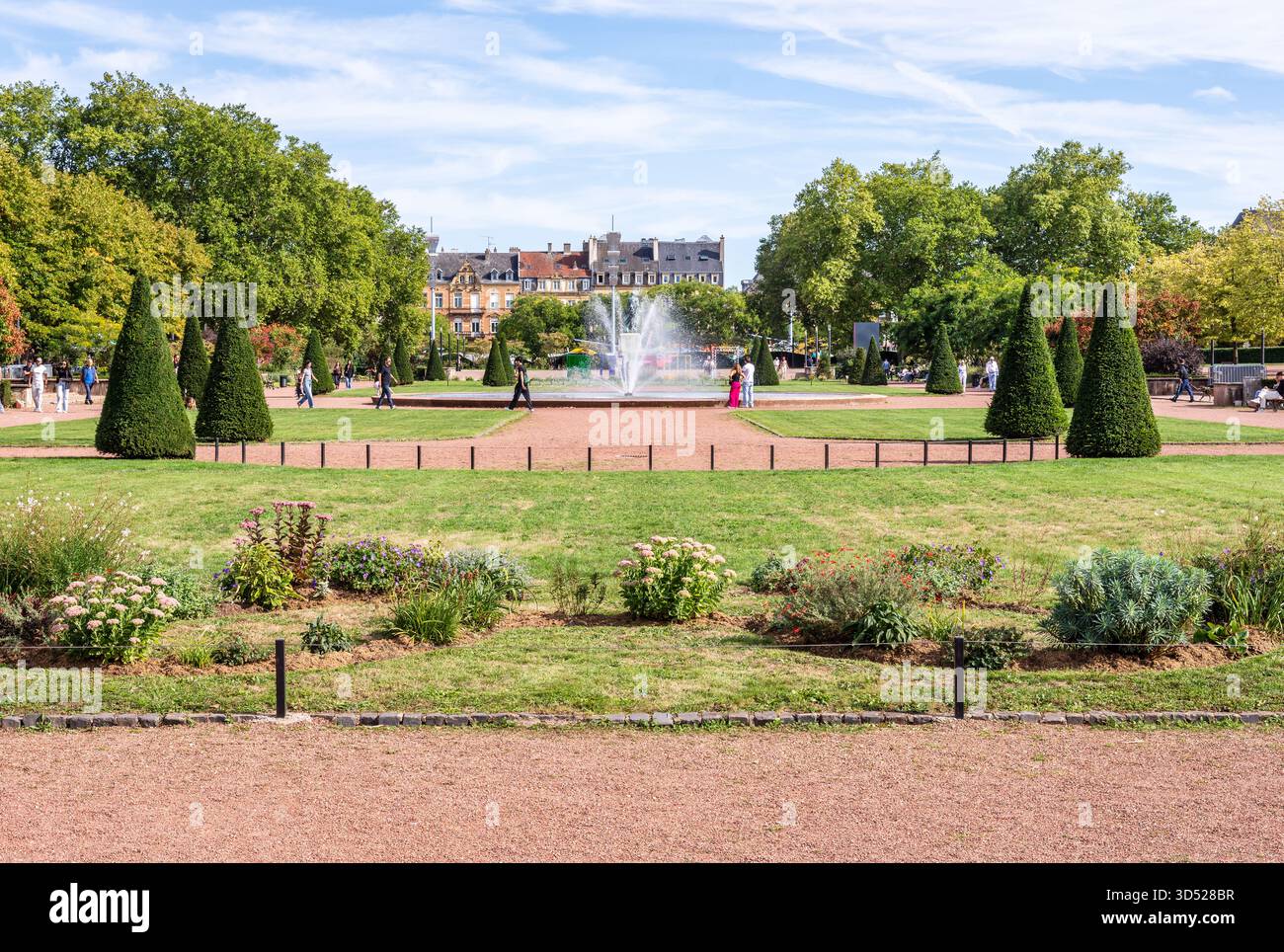 Die Menschen genießen einen sonnigen Tag im Garten der Esplanade in Metz, Frankreich, einem öffentlichen formellen Garten mit Schotterwegen, getrimmten Bäumen und einem zentralen Brunnen. Stockfoto