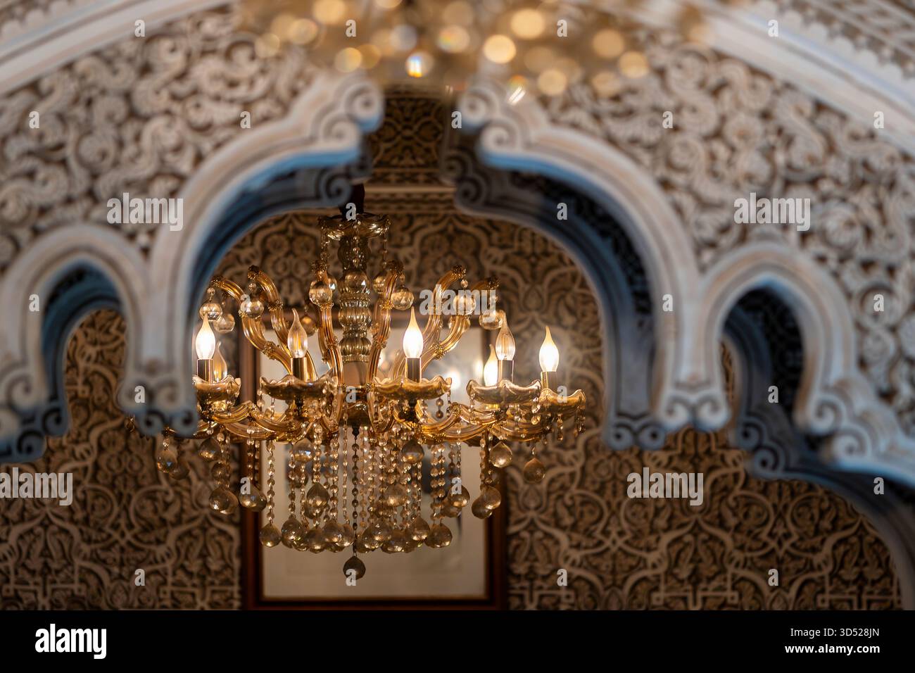 Goldener Kronleuchter, der unter kunstvollen marokkanischen Bögen mit Stuckmuster leuchtet. Eleganter Kristalllüster beleuchtet den Flur, Marrakesch. Stockfoto