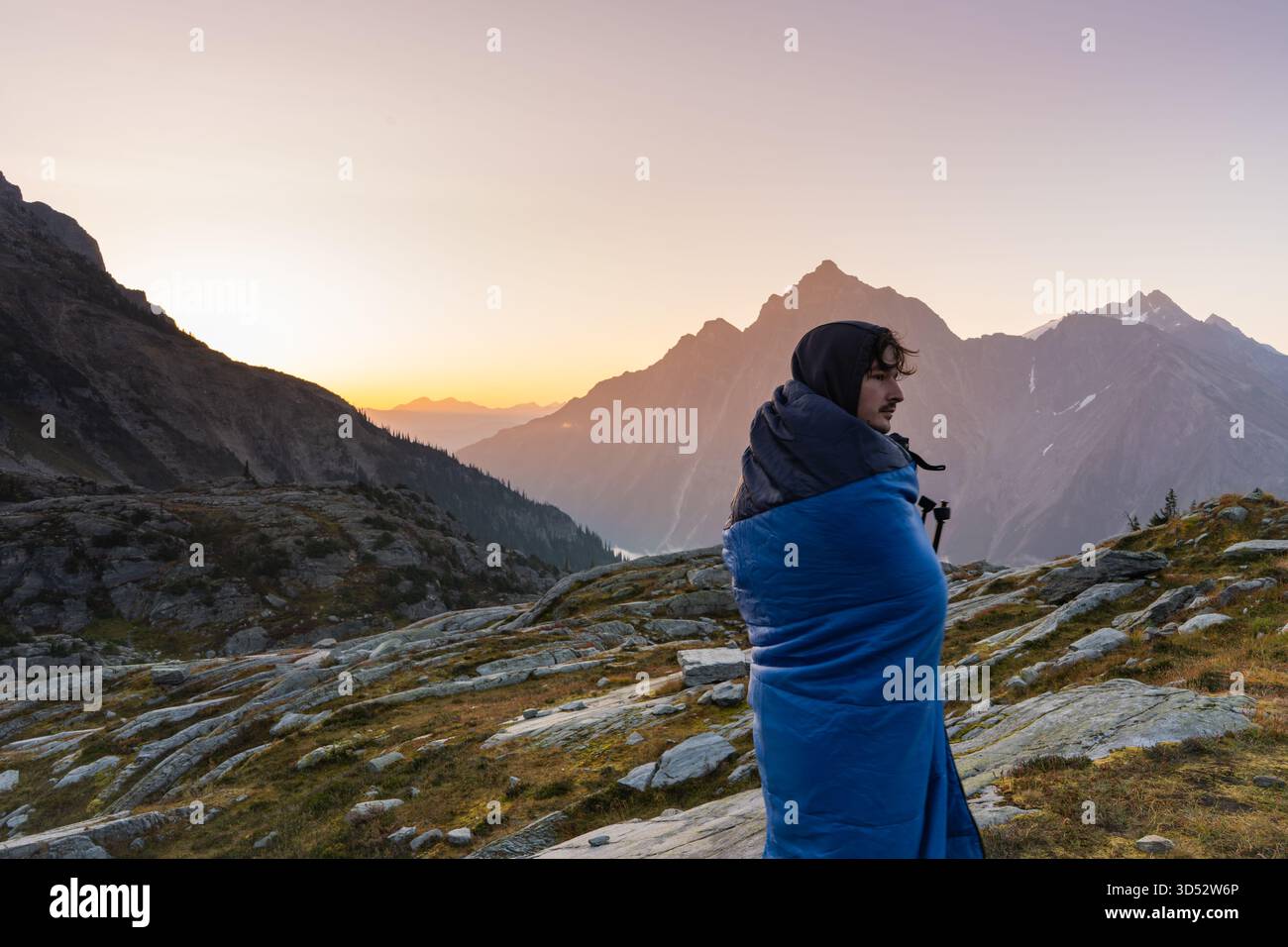 Ein Mann im Schlafsack beobachtet einen Sonnenaufgang in den Bergen. Glacier-Nationalpark, BC Stockfoto