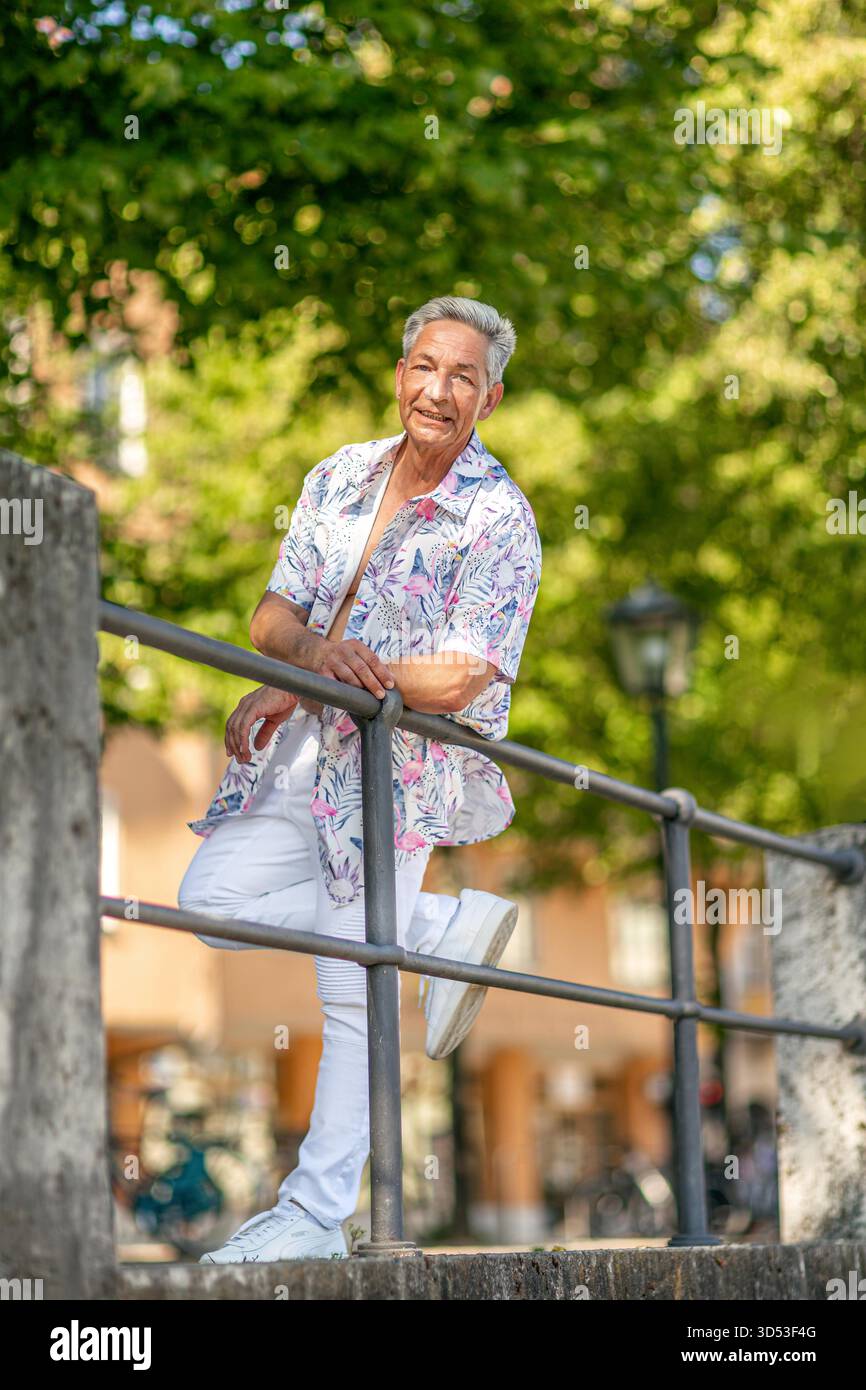 Ein fröhlicher Seniorenmann, der auf einem von Bäumen gesäumten Weg steht, ein farbenfrohes tropisches Hemd und weiße Jeans trägt. Er bewegt sich spielerisch zur Kamera und strahlt an einem sonnigen Tag in einer parkähnlichen Umgebung Selbstvertrauen und Humor aus. || Modell freigegeben Stockfoto