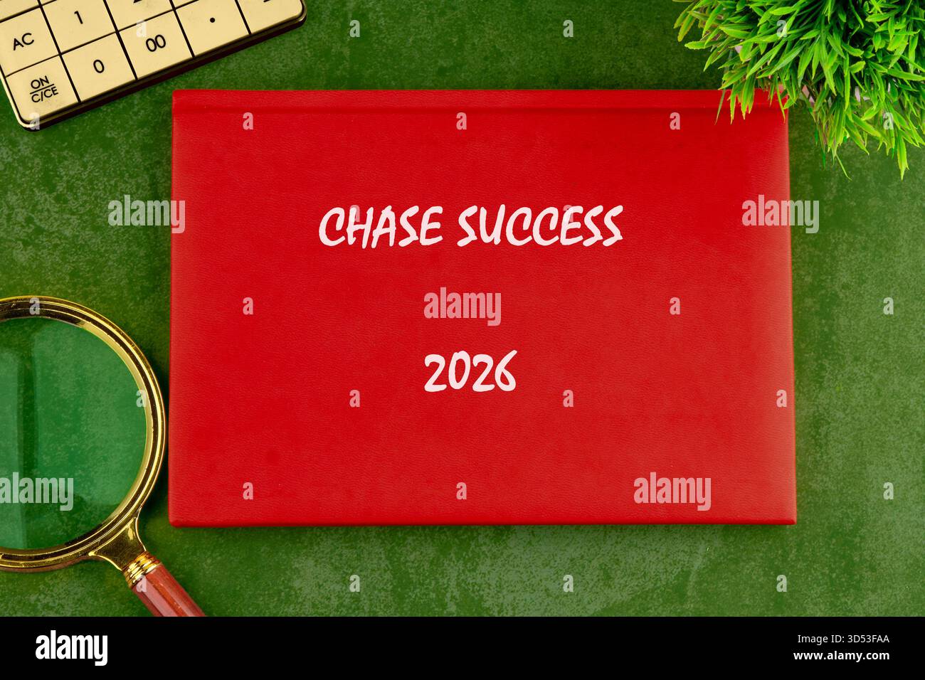 Auf einer grünen Oberfläche befindet sich ein rotes Buch mit der Bezeichnung CHASE SUCCESS 2026 neben einem Rechner, einer Lupe und einer grünen Pflanze, was Planung und Fokus anzeigt. Stockfoto
