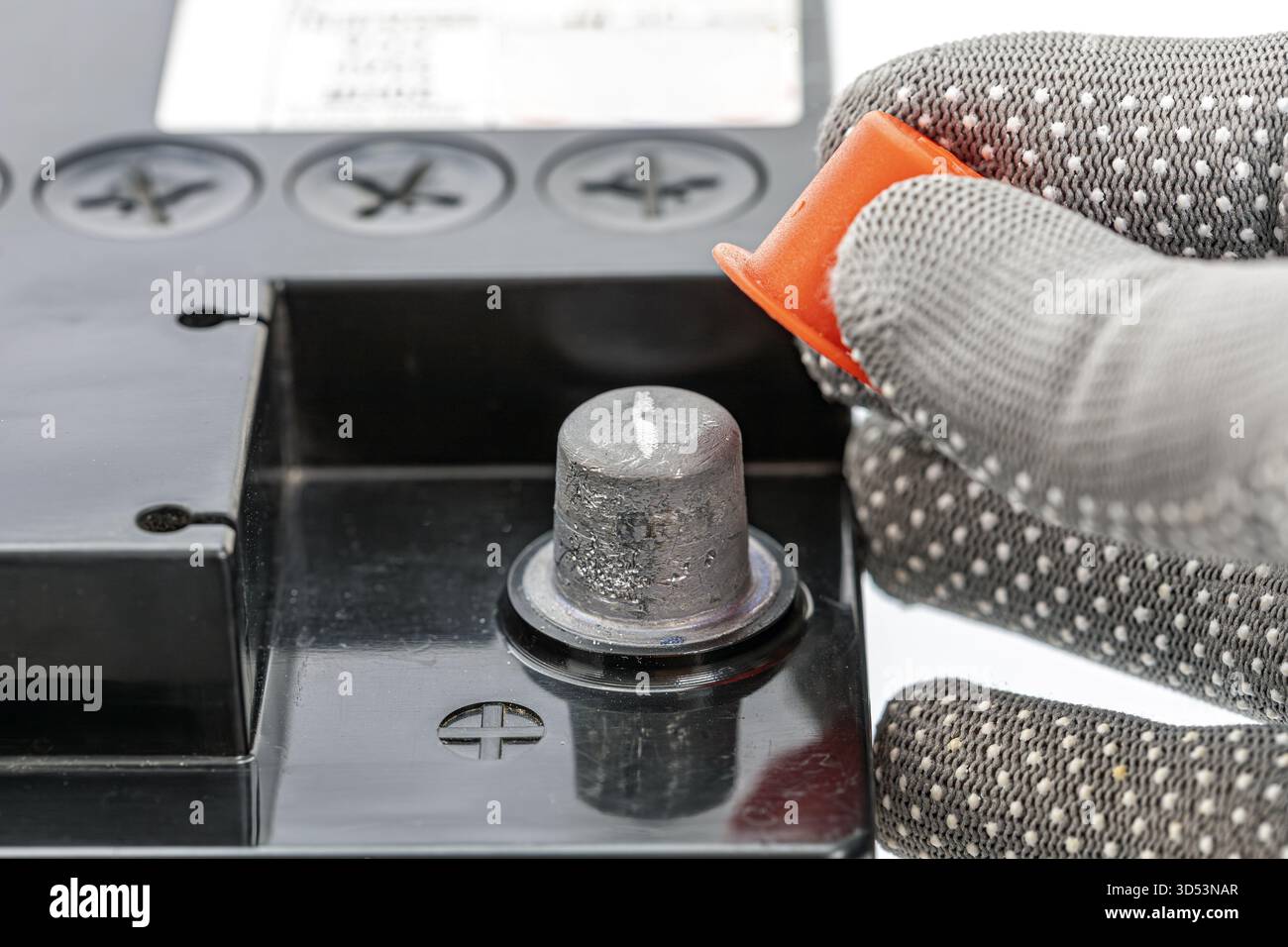 Eine Hand mit Handschuhen löst und entfernt die hellorangefarbene Schutzkappe von einem Autobatteriepol, sodass der darunter liegende Metallanschluss freiliegt. Das Batteriefach Stockfoto