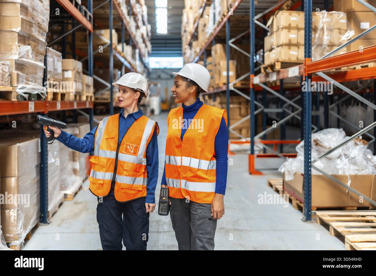 Zwei Frauen, die Schutzhelme und Sicherheitswesten tragen, analysieren den Bestand, indem sie Produktbarcodes in einem modernen Logistiklager scannen Stockfoto